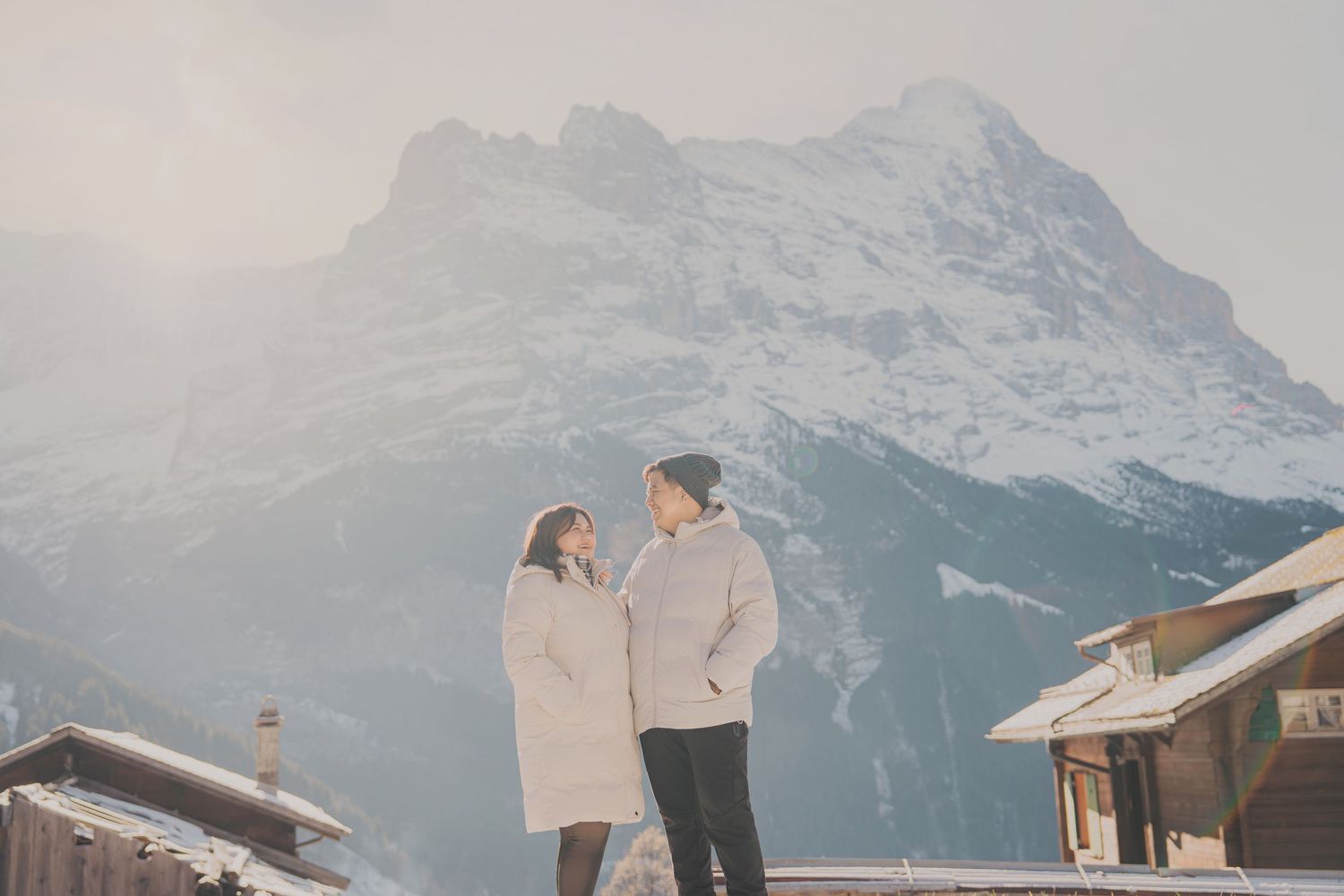 Romantic couple in white winter coats gazing at each other with misty snowy mountain backdrop.