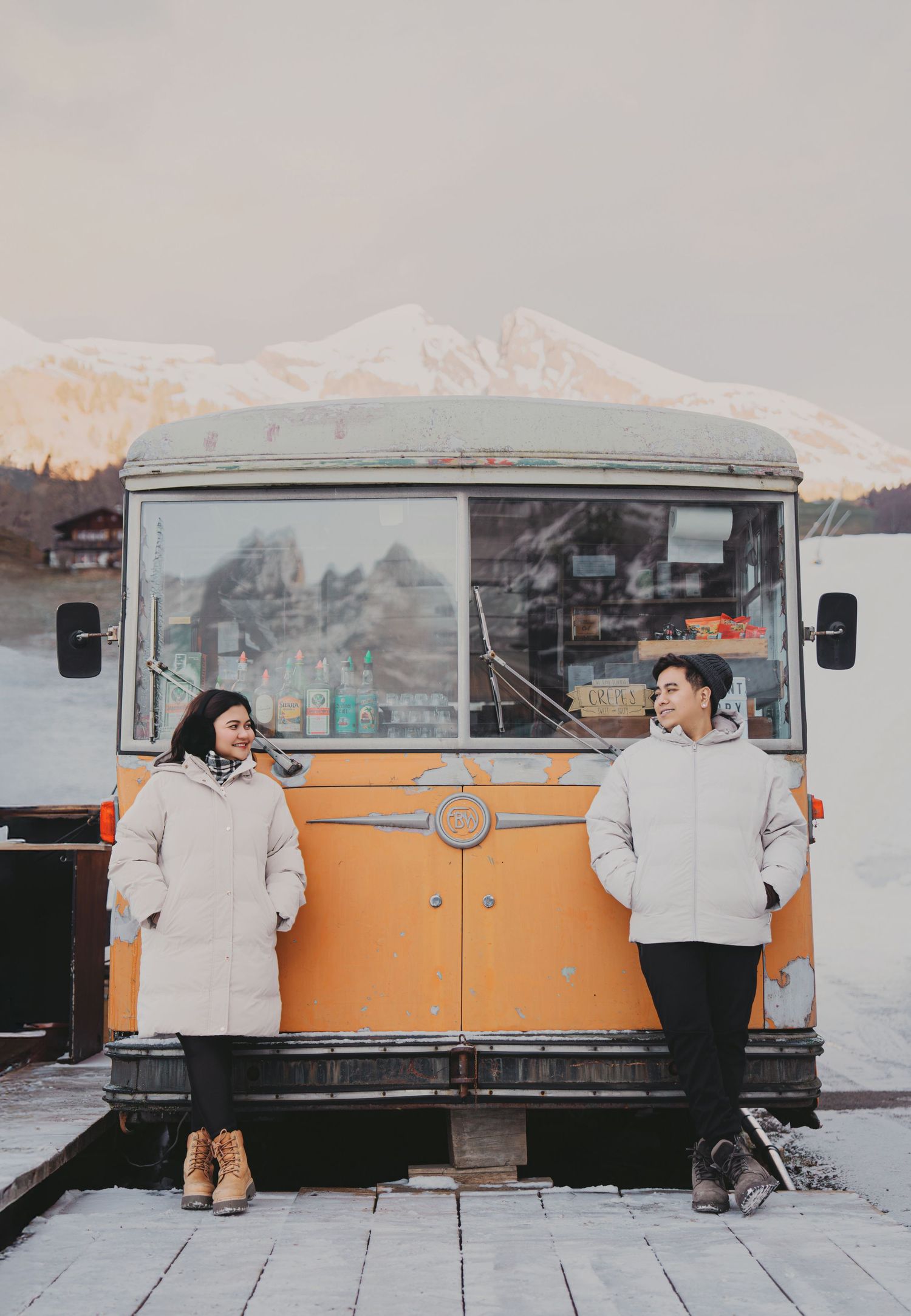 Couple in matching white puffer jackets leaning against a vintage orange food truck in a snowy mountain setting.