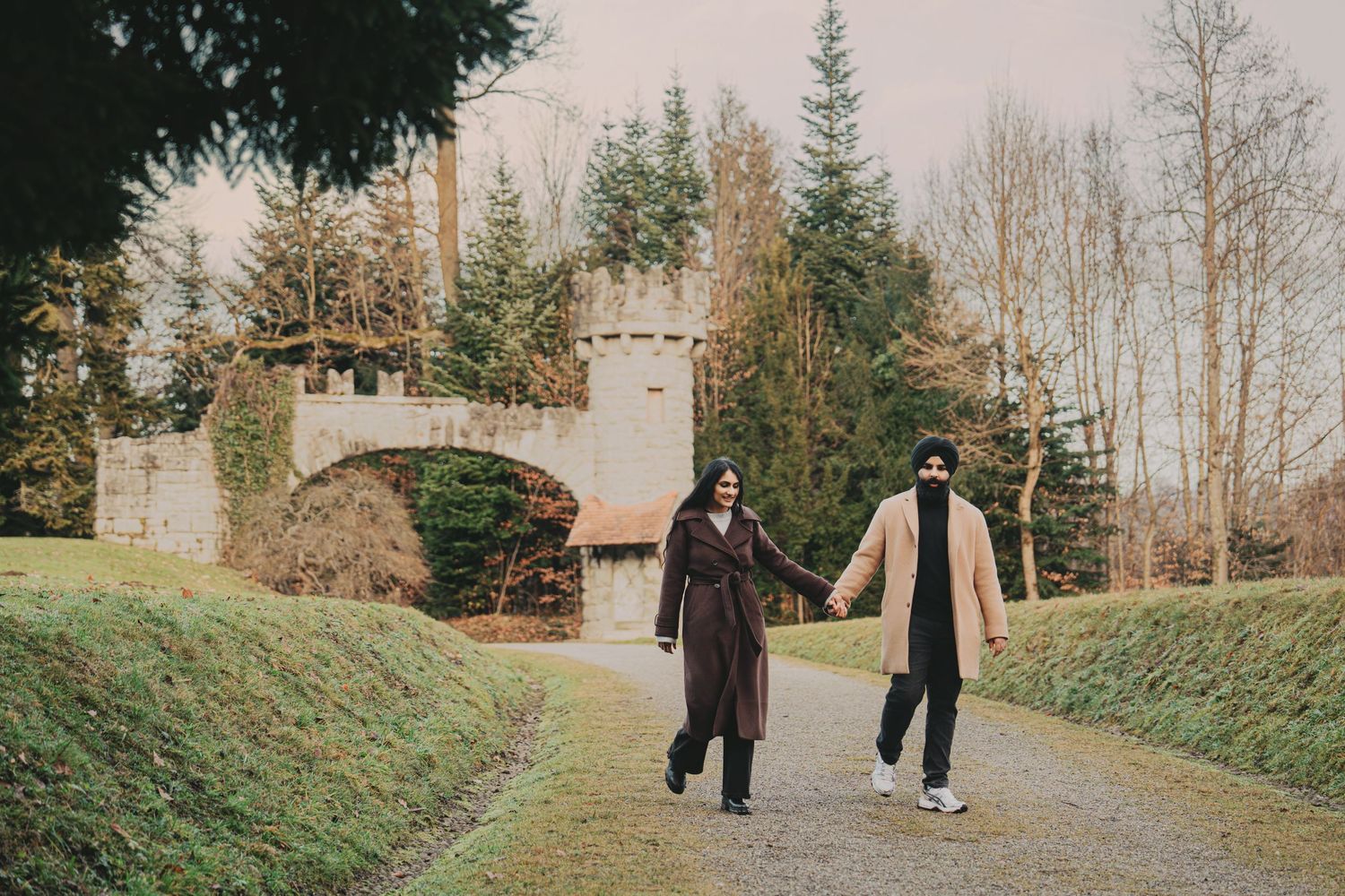 Couple walking hand in hand on a pathway with a stone castle arch bridge behind them in winter.
