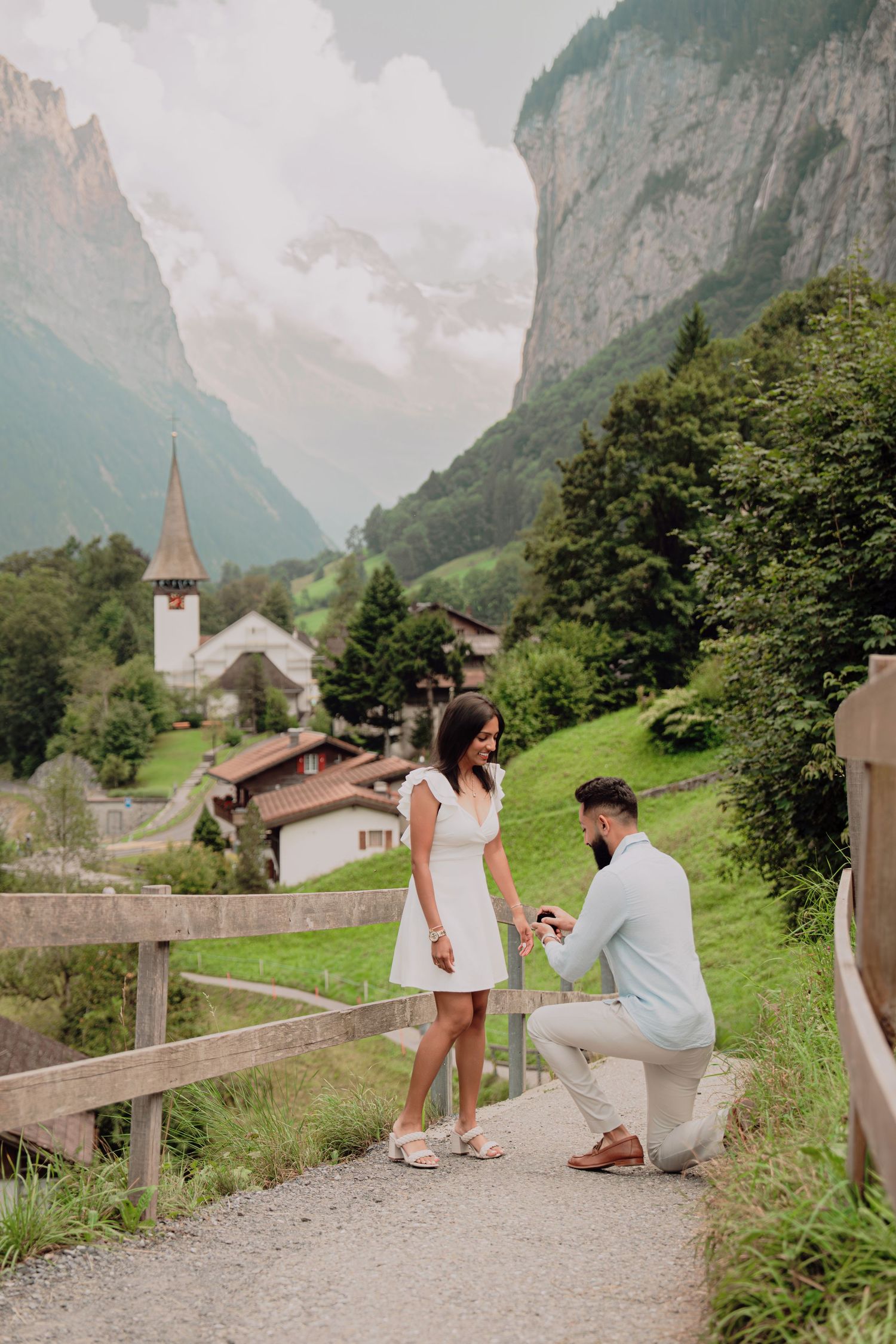 A man kneels on a mountain path presenting a ring to a woman with a picturesque Swiss Alpine village backdrop.