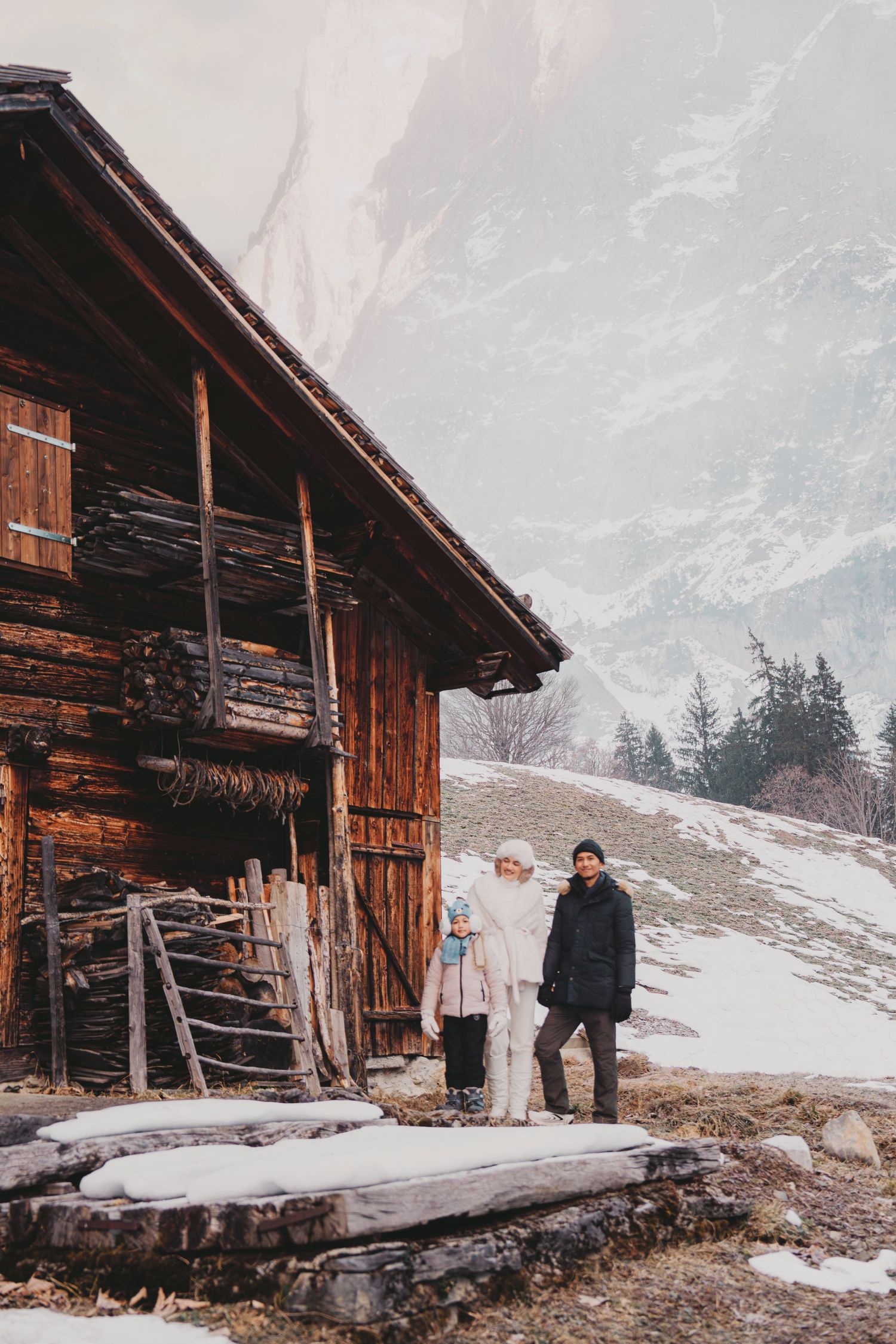 Family posing beside a weathered wooden mountain barn with snowy peaks in the background.