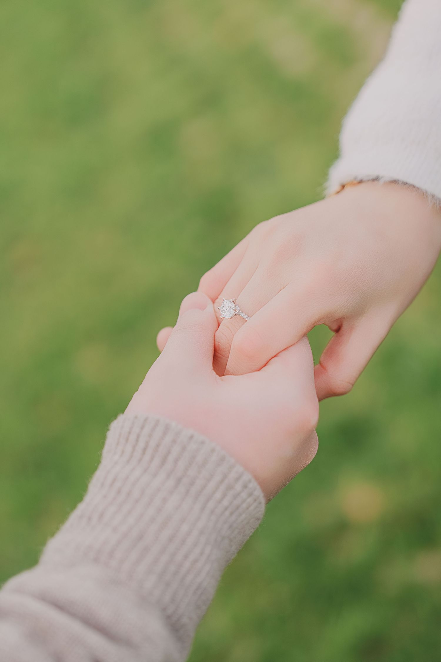 Close-up of hands holding showing diamond engagement ring on green grass background.