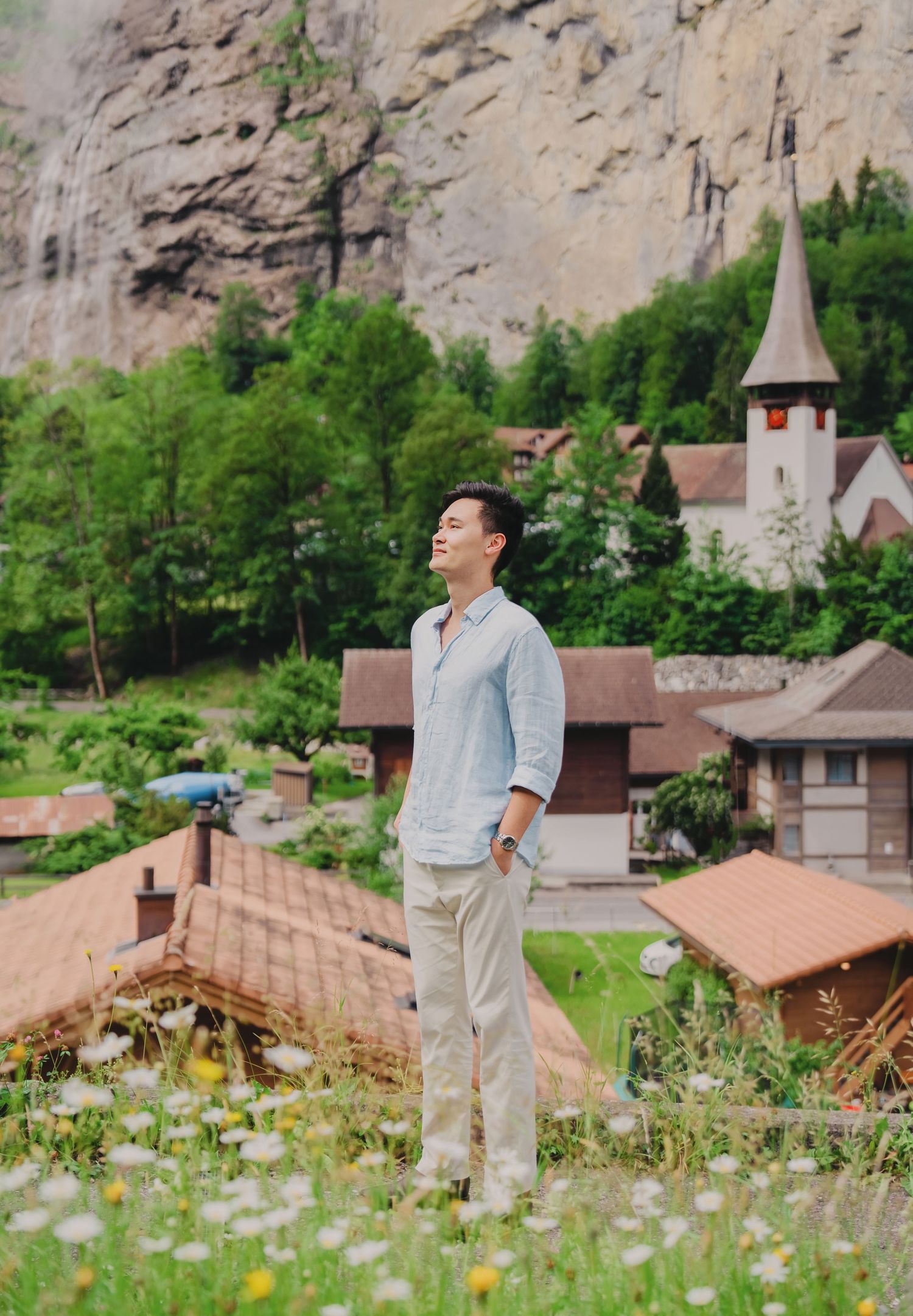 Man standing in flower field with Swiss village church and rocky mountain waterfall backdrop.