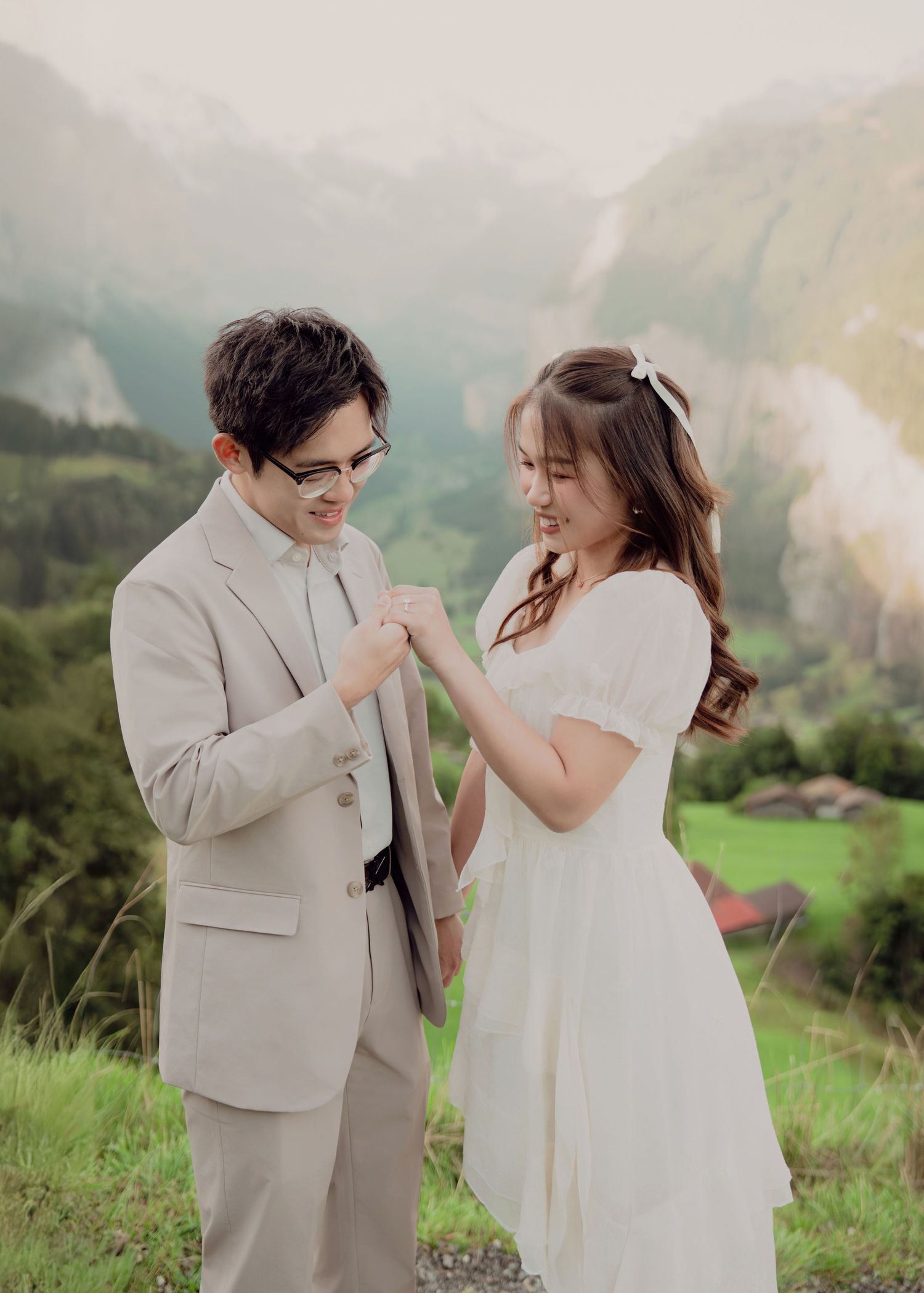 Smiling couple in wedding attire exchanging rings on Swiss alpine meadow with misty mountain valley backdrop.
