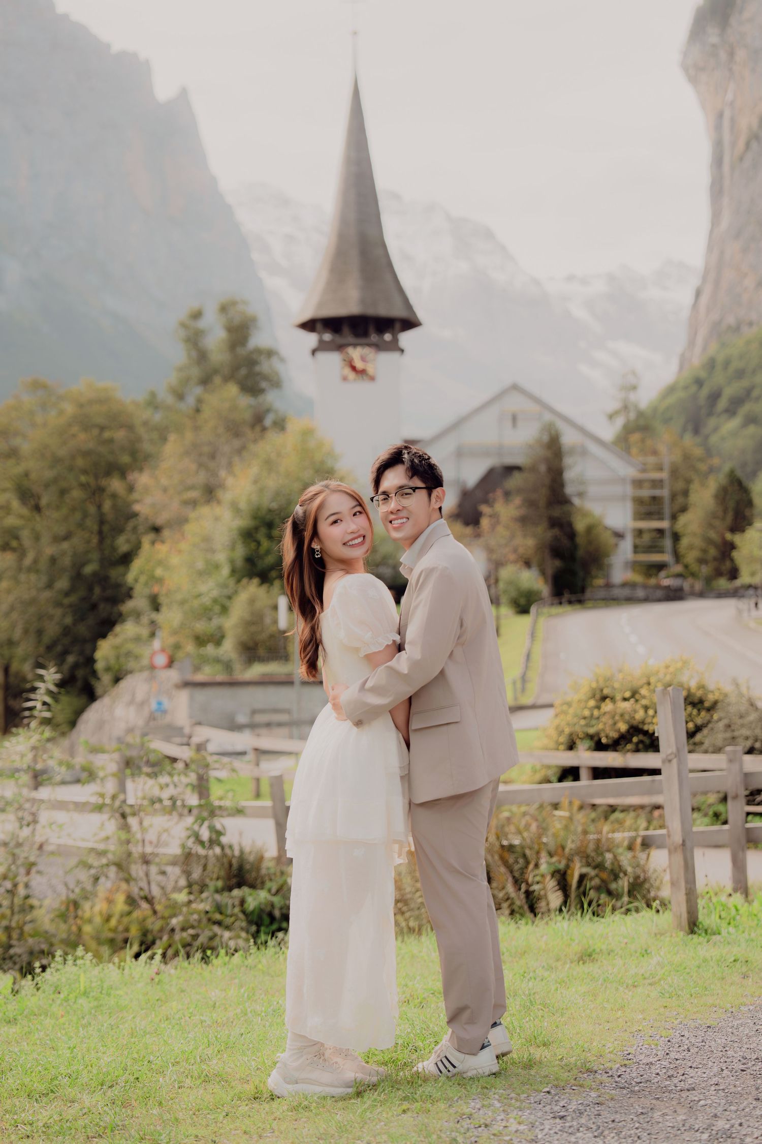 Smiling couple embracing in front of a historic Swiss church with mountain backdrop.