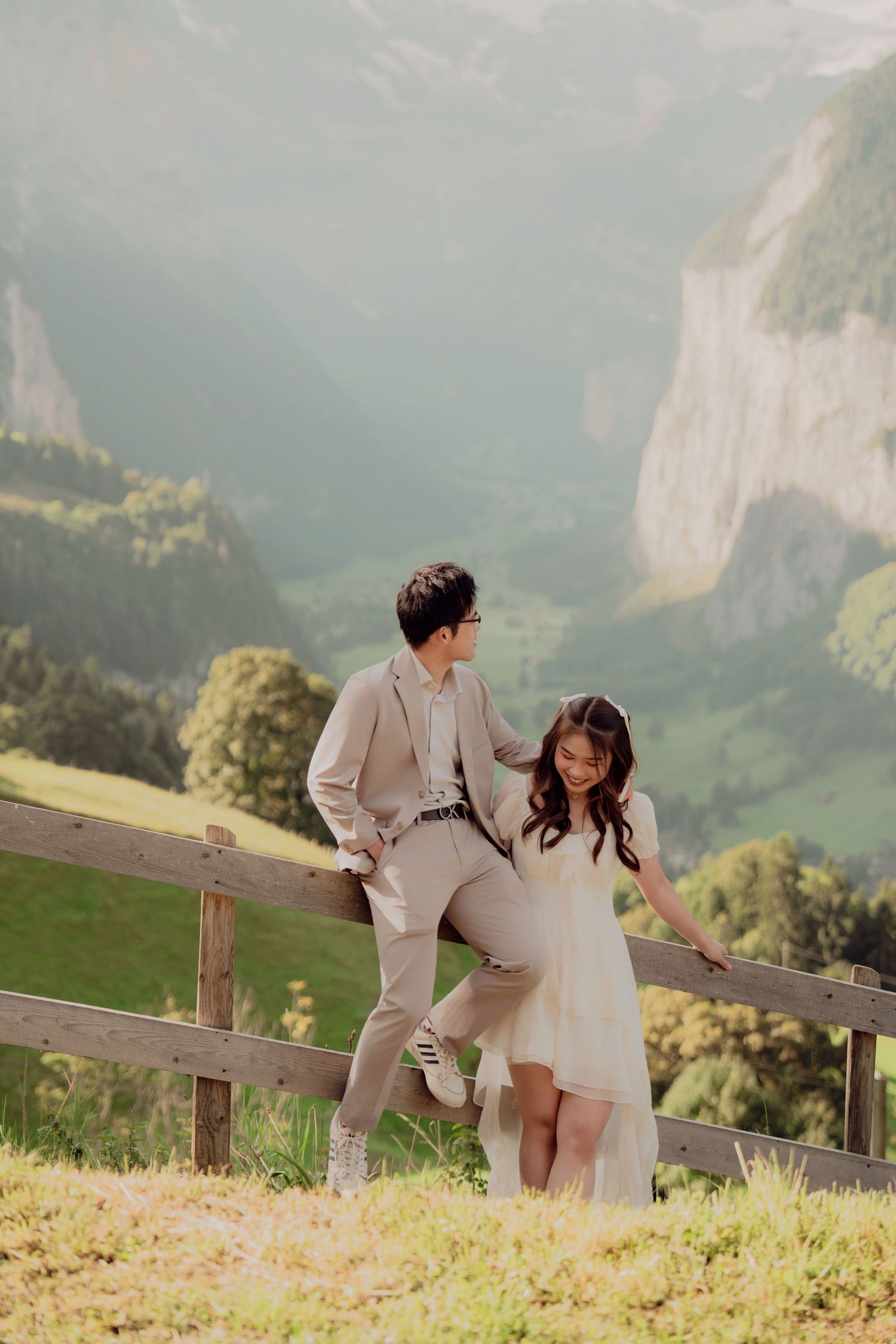 Elegant couple posing by a wooden fence with dramatic Swiss alpine valley and cliffs behind them.