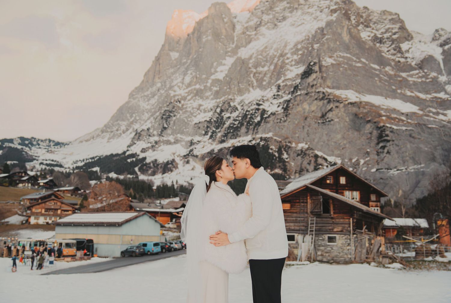 Couple sharing a kiss in matching white outfits with snowy Alpine village and mountain scenery behind.