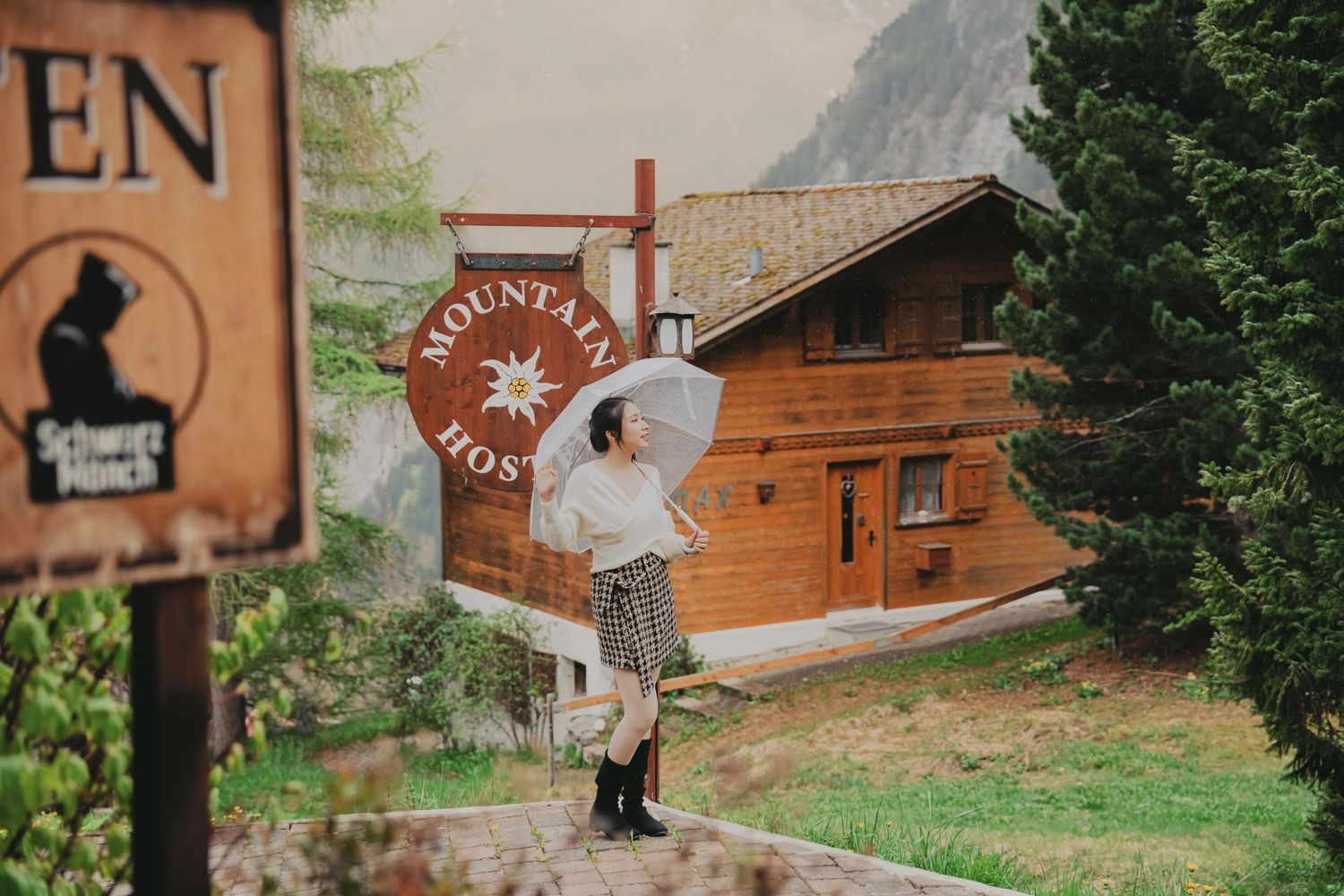 Woman posing with white umbrella near Mountain Hostel sign beside log cabin in alpine setting.