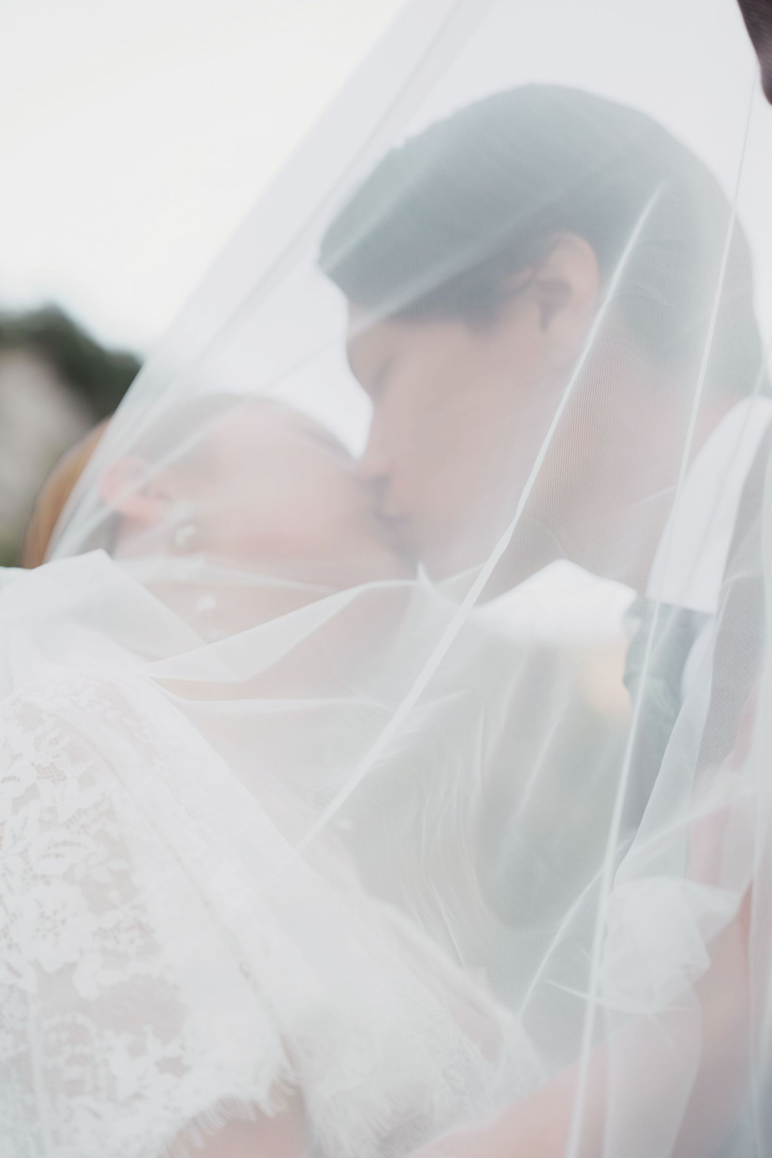 Bride and groom sharing a romantic kiss under a soft white wedding veil in an ethereal close-up portrait.
