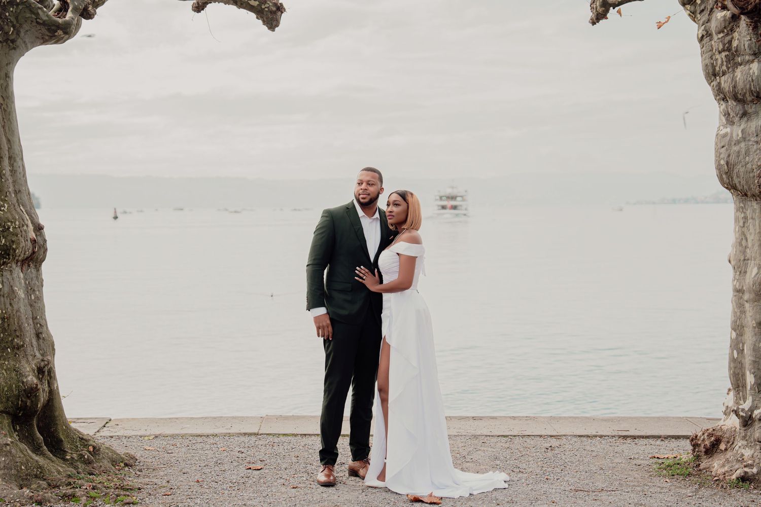 Bride and groom embracing between ancient trees with calm lake and overcast sky in background.