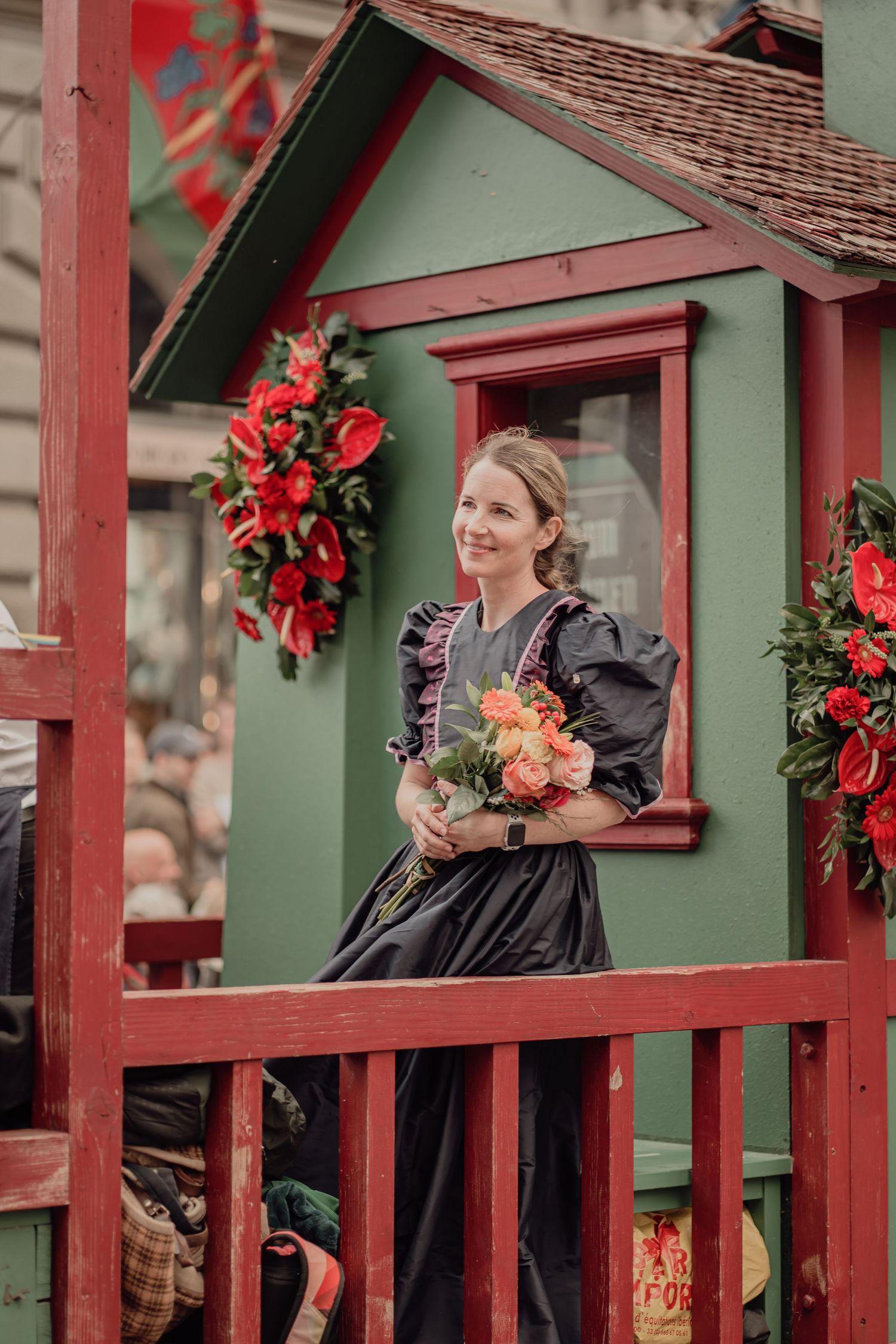 A woman in a black Victorian dress holds a bouquet of pink flowers on a decorated red and green float.