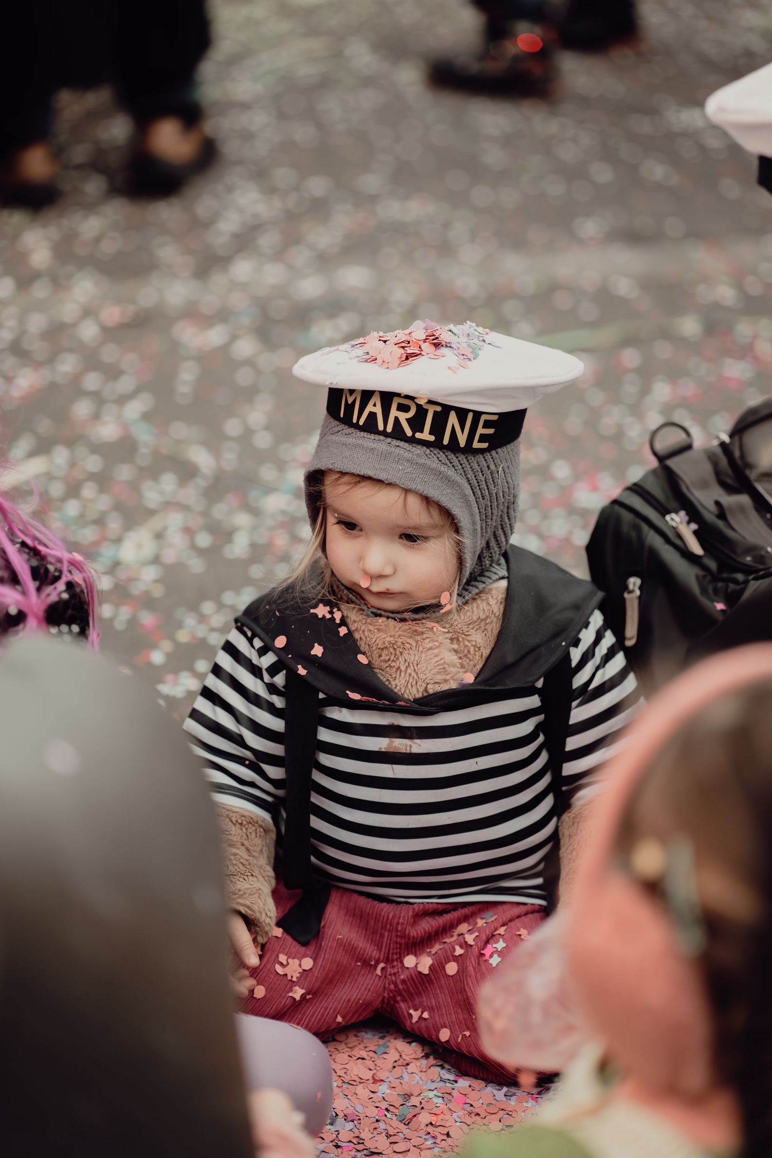 Toddler in sailor costume with Marine hat sits among colorful confetti at a carnival parade.