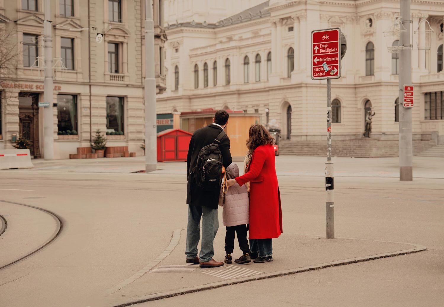 Family viewed from behind holding hands on a city tram square with neoclassical building.