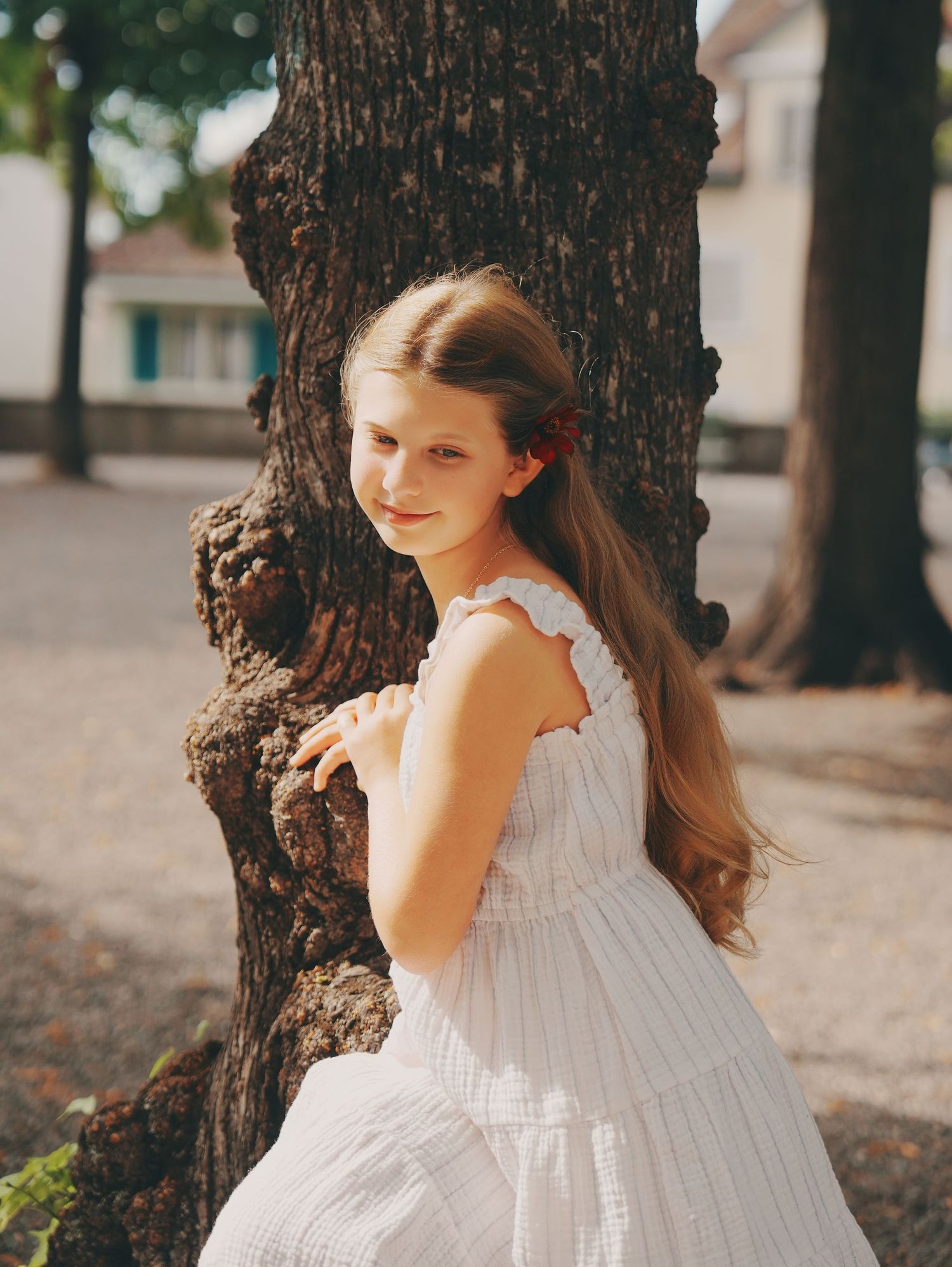 A young girl with long hair in a white ruffled dress poses beside a large tree in warm sunlight.