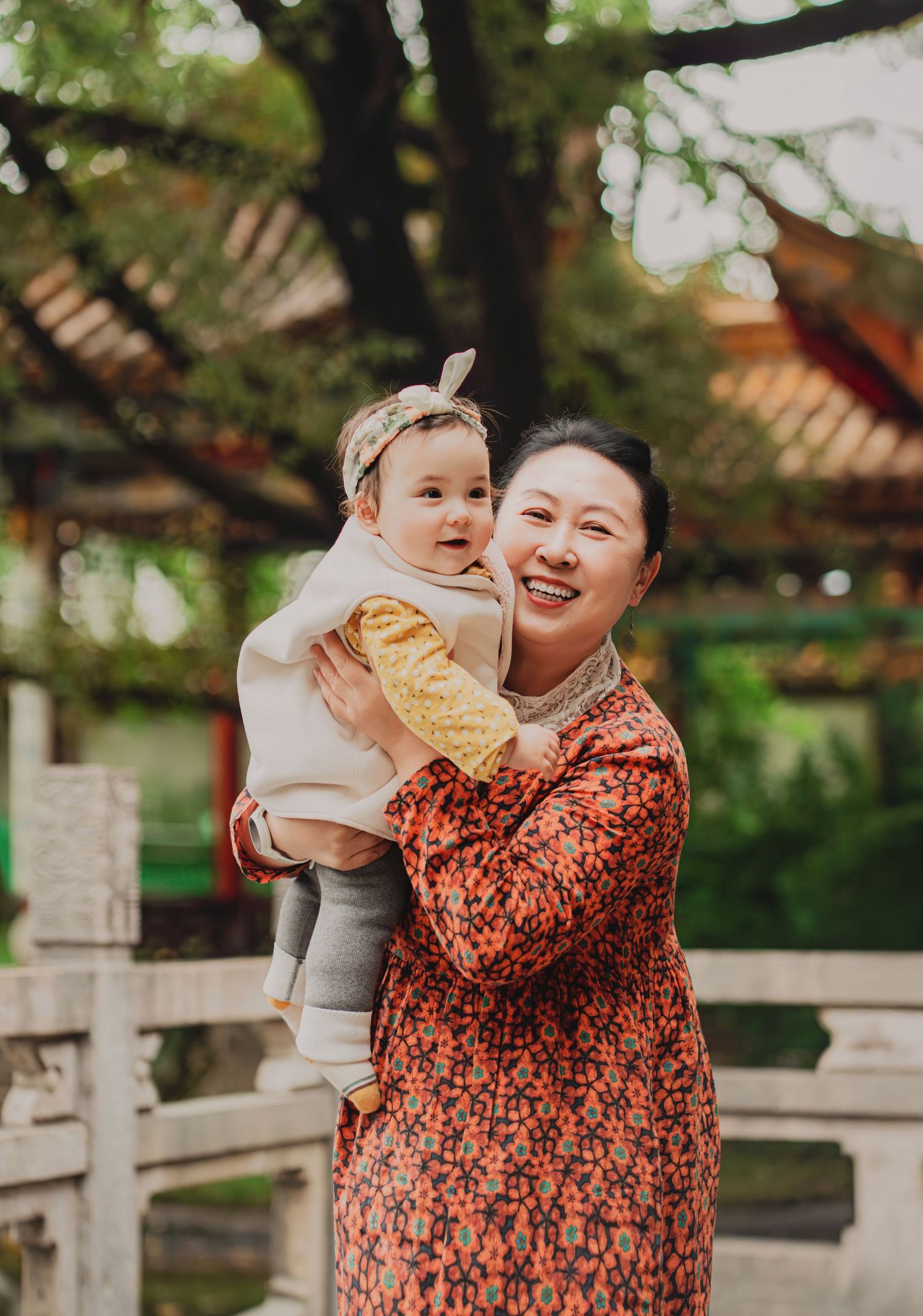 Smiling Asian grandmother holding adorable baby with headband in traditional Chinese garden setting.