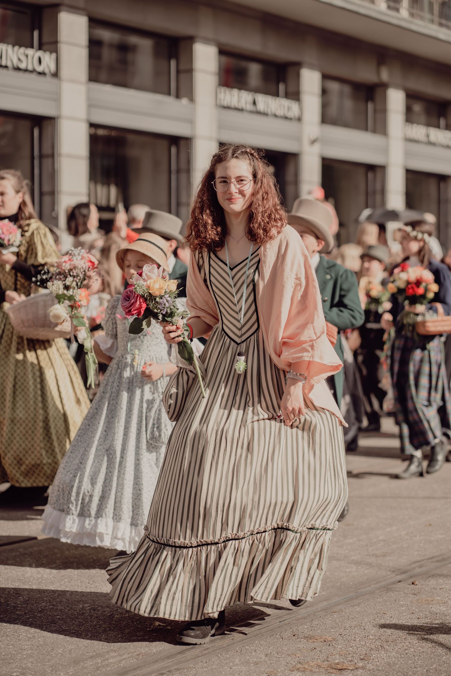 Smiling young woman in a striped historical gown holds flowers during a sunny outdoor parade.