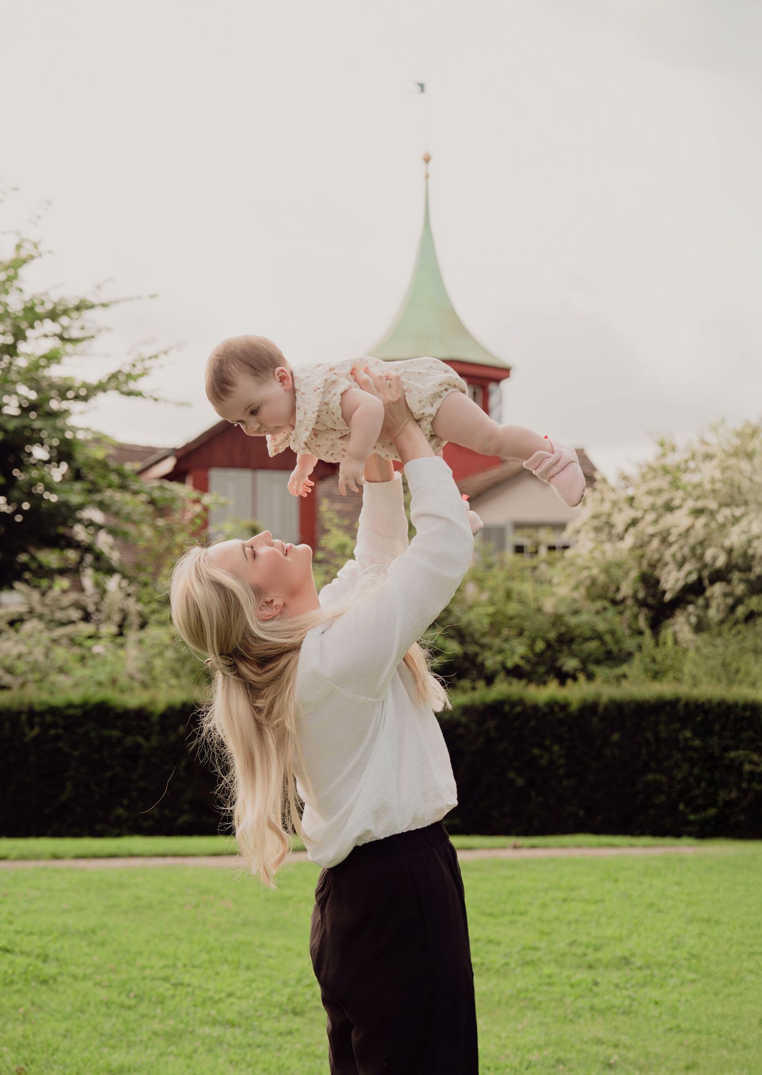 Blonde mother lifting smiling baby high in the air in a green park with a red-roofed tower in background.