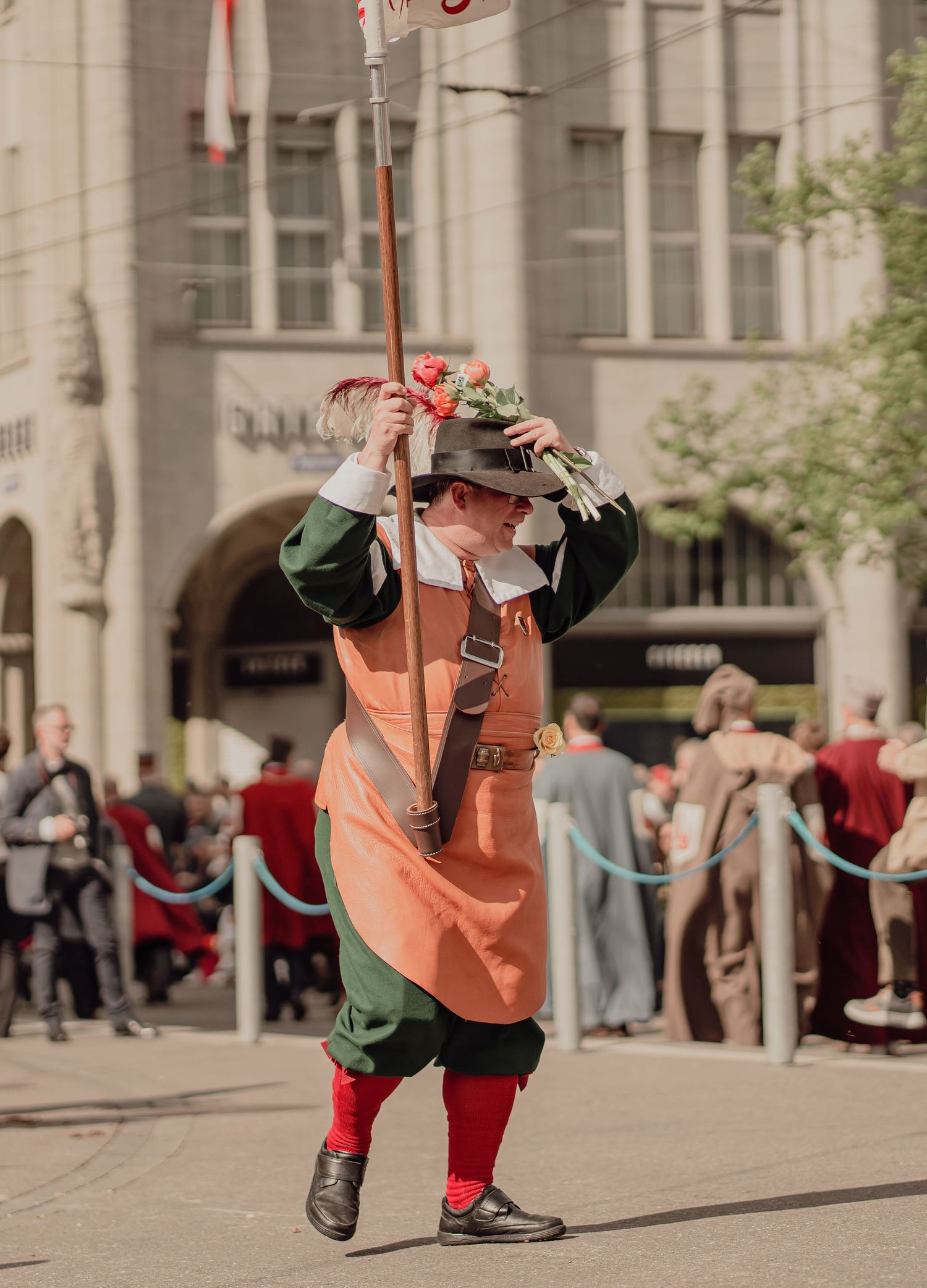A performer in orange and green historical costume twirls a tall flag-pole during a street parade.