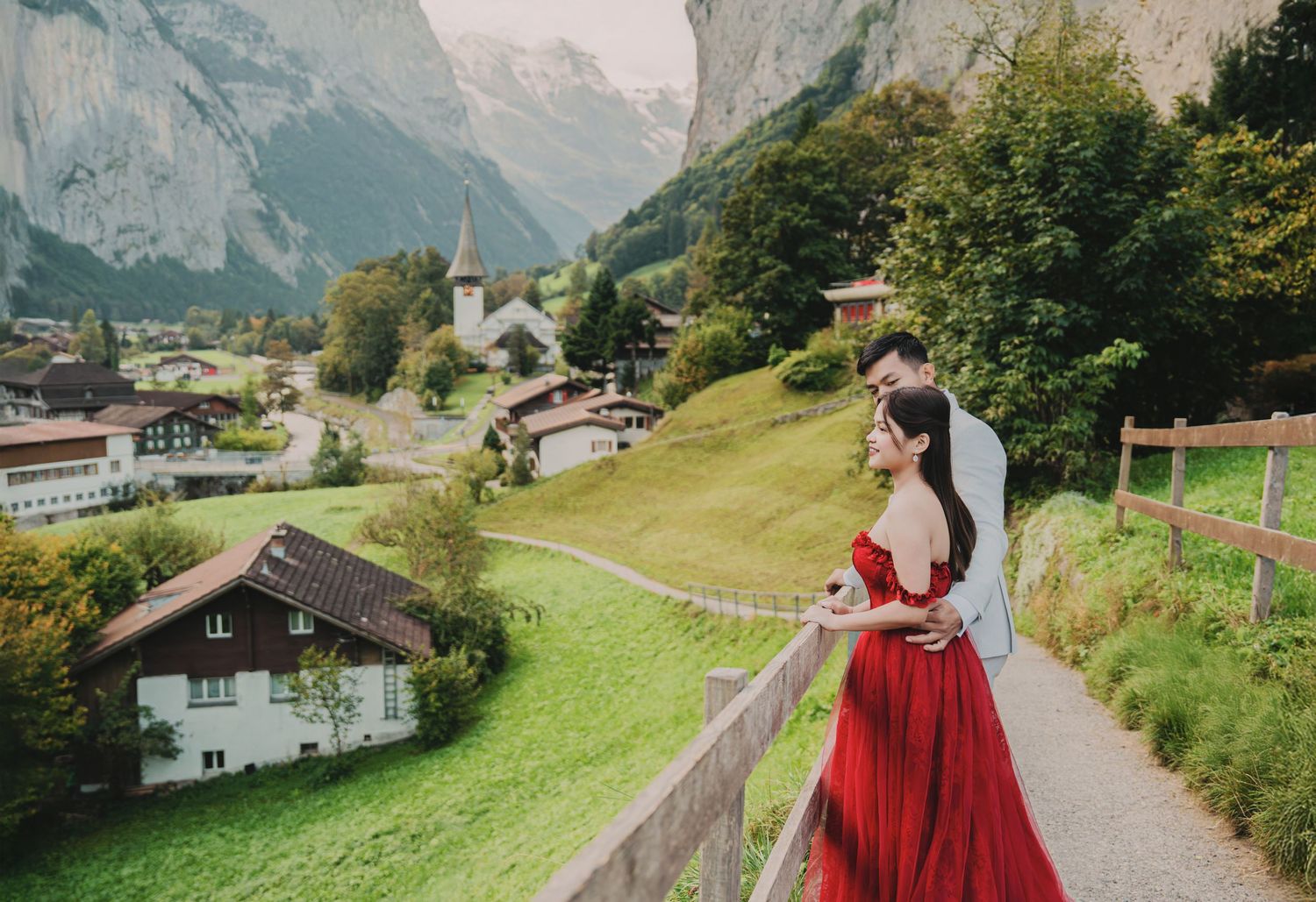 Couple gazing at scenic Swiss alpine village, woman in red off-shoulder gown, man in light grey suit.