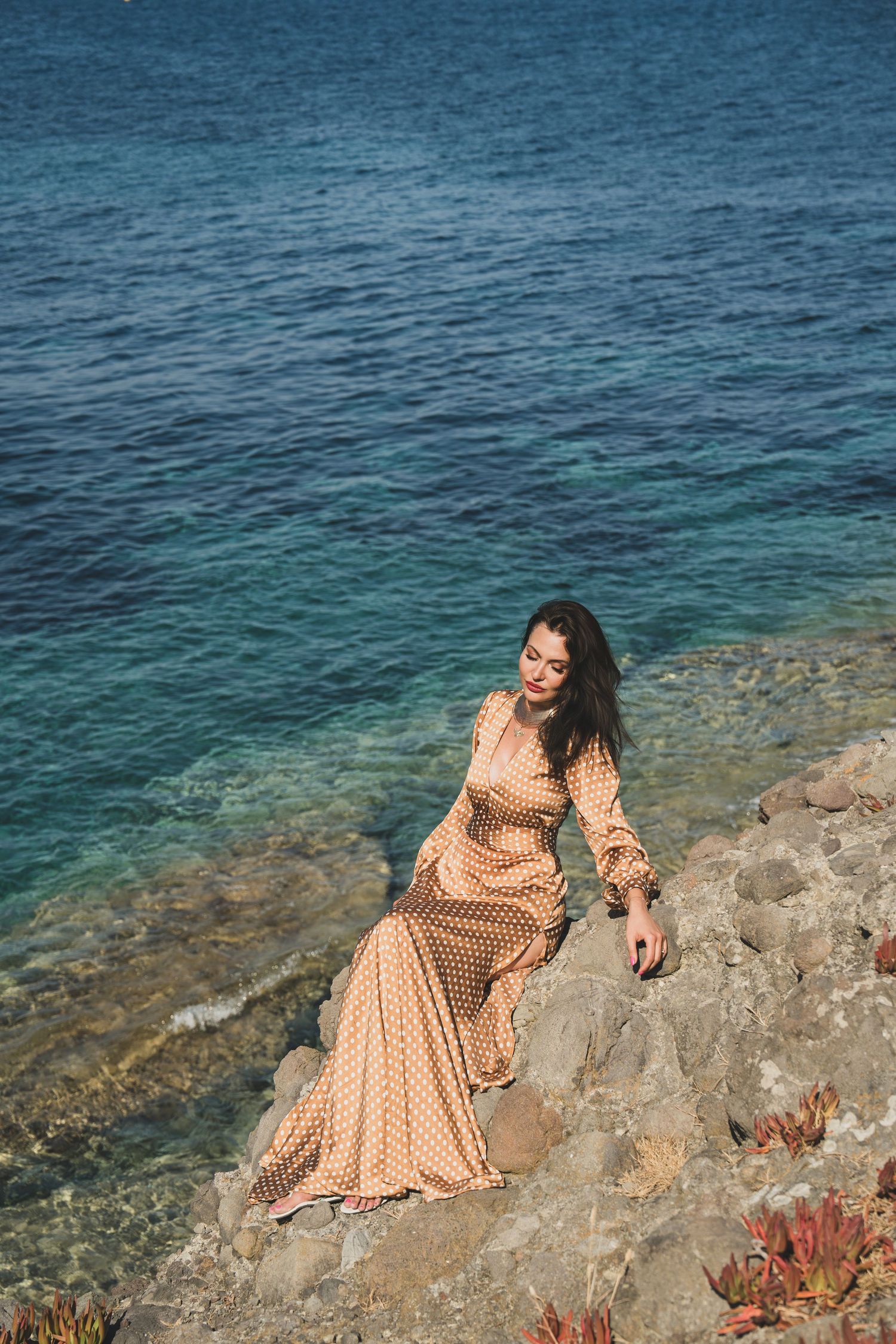 Woman in long polka dot dress sits on coastal rocks beside clear turquoise Mediterranean sea.