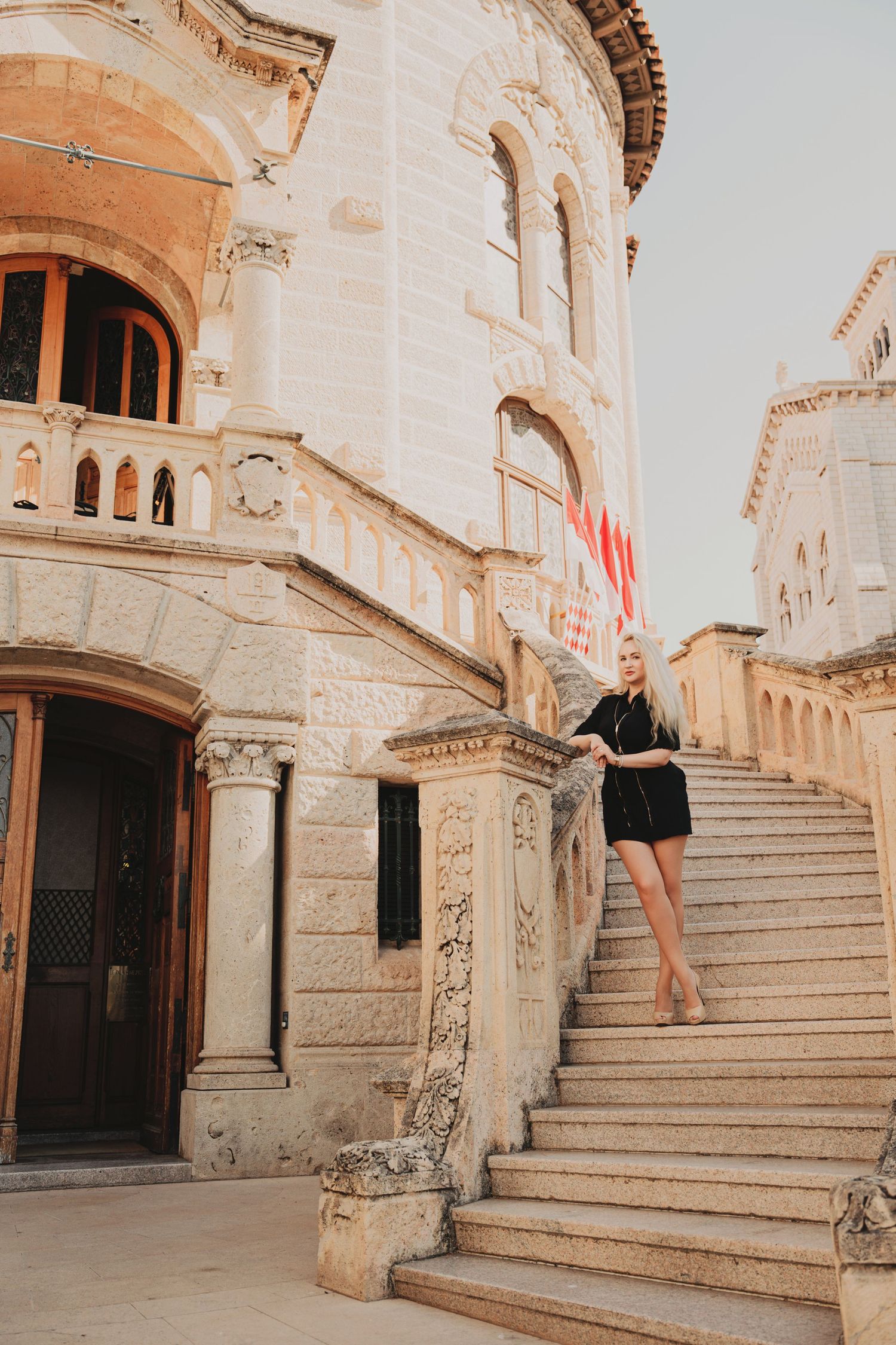 Blonde woman in black dress posing on grand stone steps of Monaco courthouse.