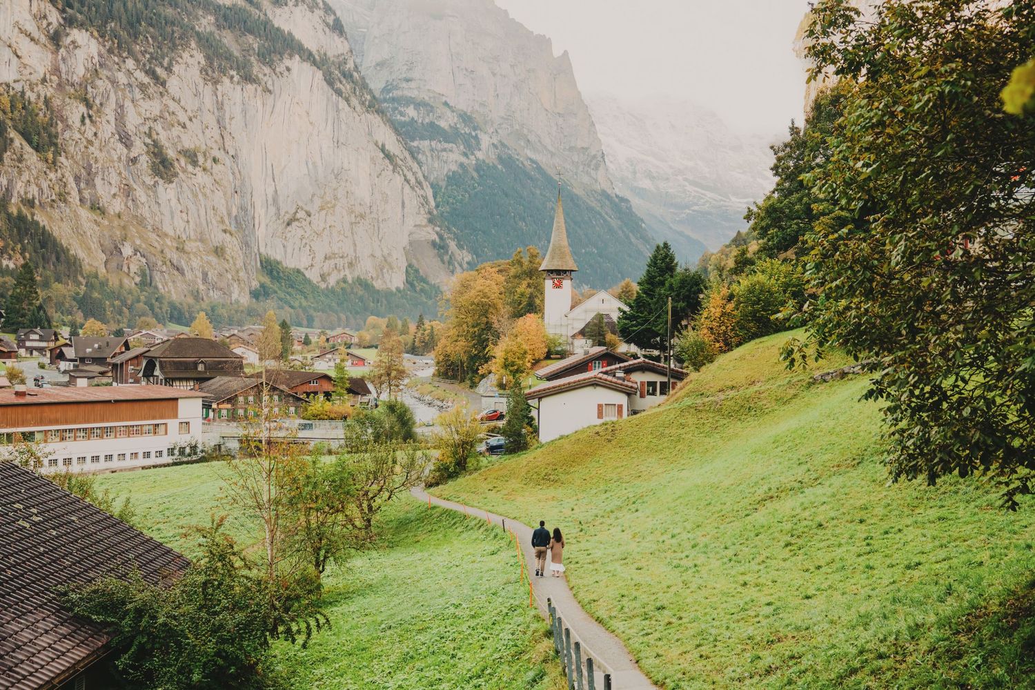 Two people strolling down a fenced path toward a charming Swiss Alpine village with towering mountains behind.