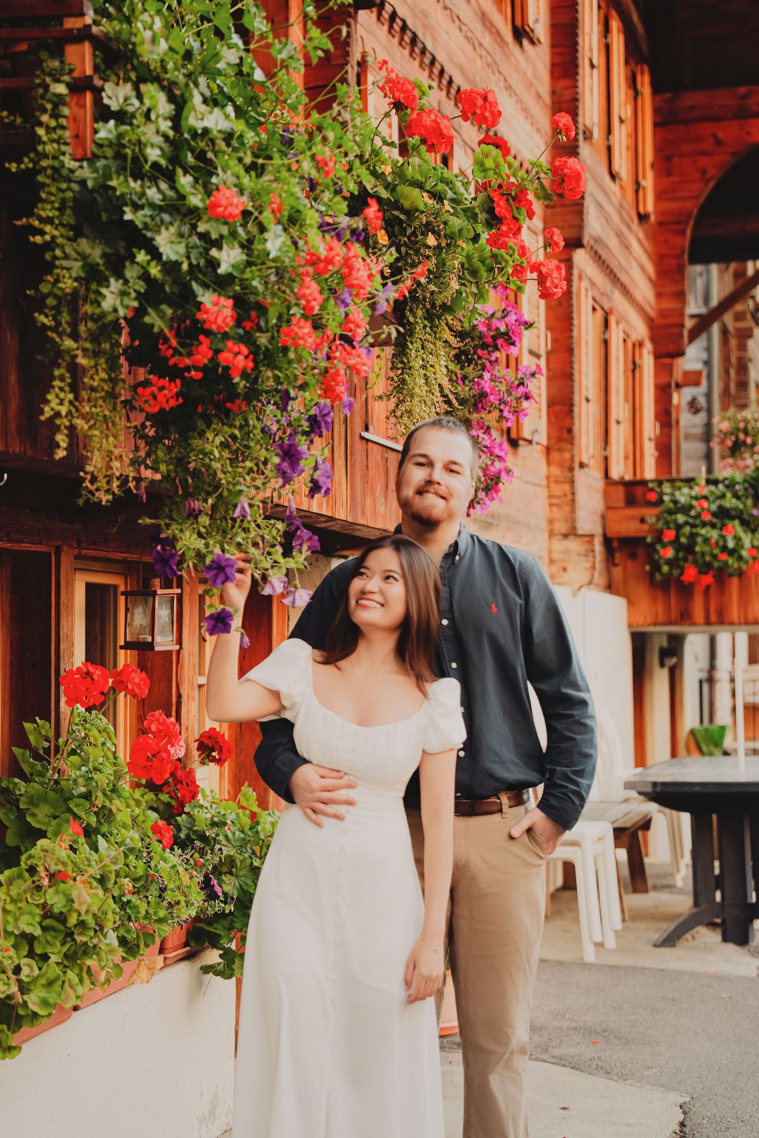 Happy couple embracing in front of a rustic wooden building with vibrant red and purple flowers.