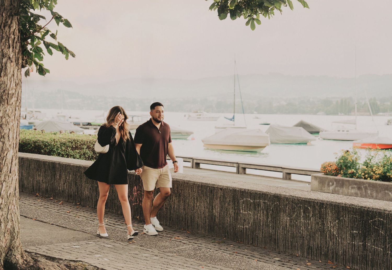 Couple strolling together on a waterfront path with moored boats and misty mountains behind them.