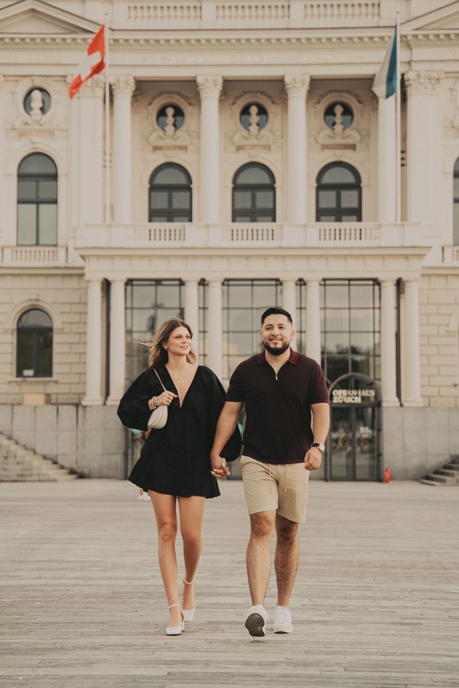 Couple strolling hand in hand across a plaza with a grand white European building behind them.