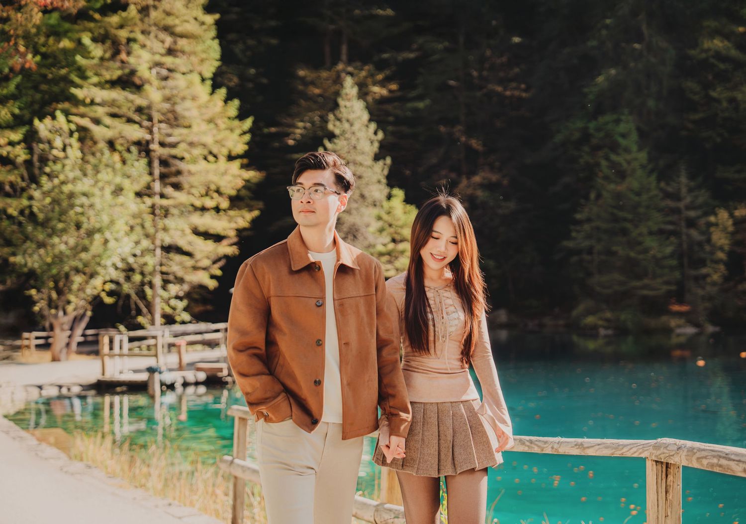 Young Asian couple holding hands walking by turquoise lake in autumn forest setting.