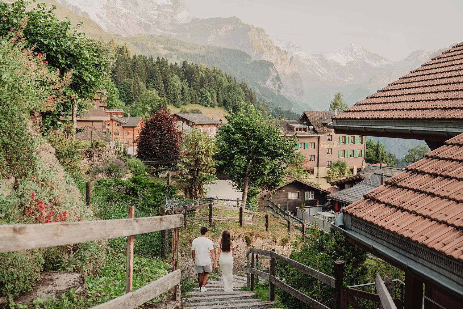 Couple walking hand in hand down wooden stairs through lush Swiss village with Alpine mountain views.