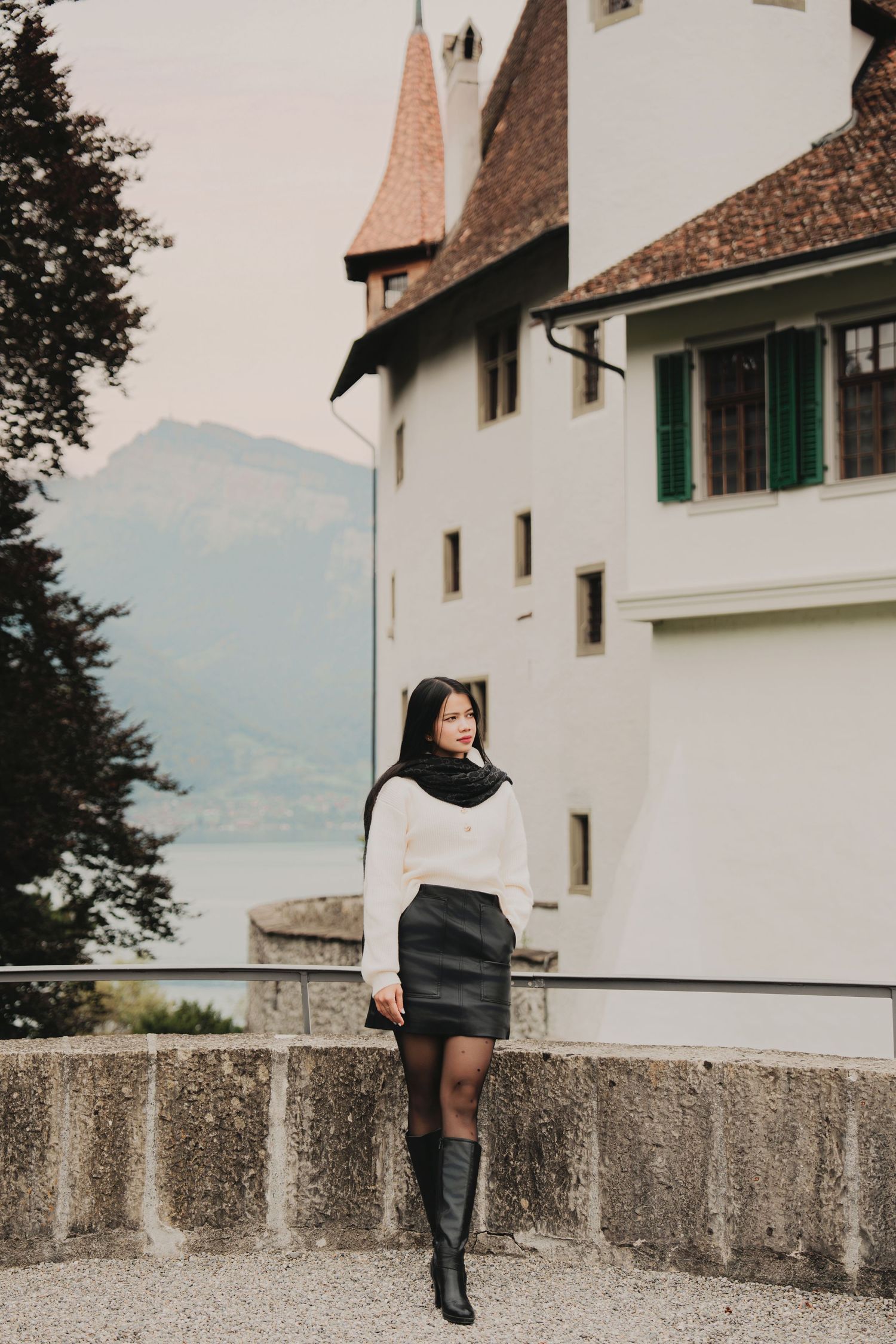 Young woman in white sweater and black leather skirt poses before a white Swiss castle tower.