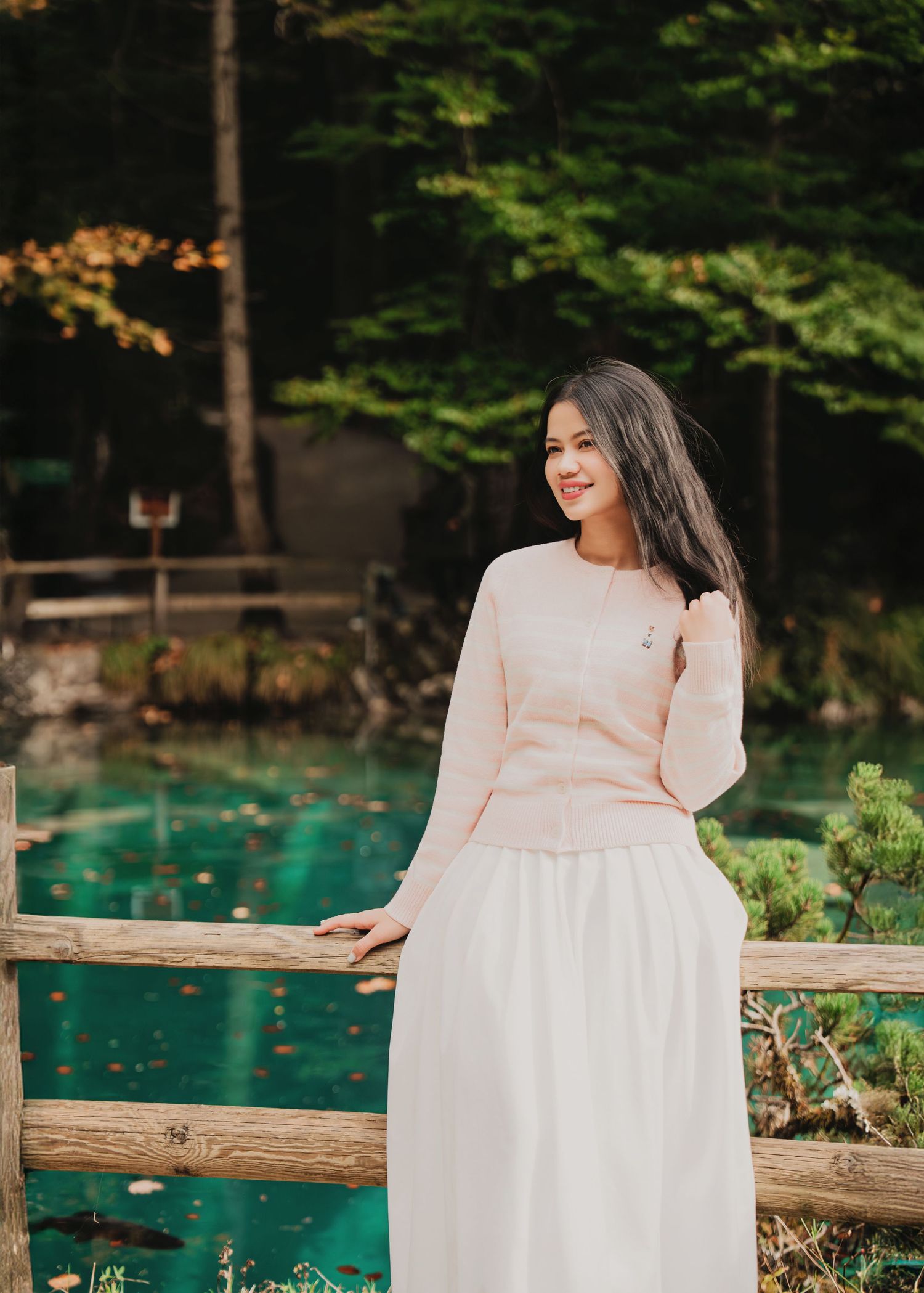 Young woman in pink top and white skirt smiling by turquoise lake with wooden fence and forest backdrop.