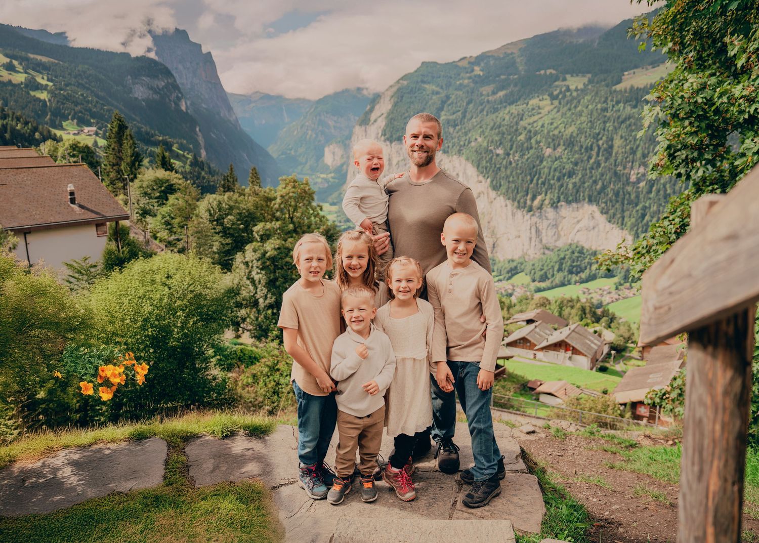 Father holding baby surrounded by six children posing on path with dramatic Swiss Alps valley view.