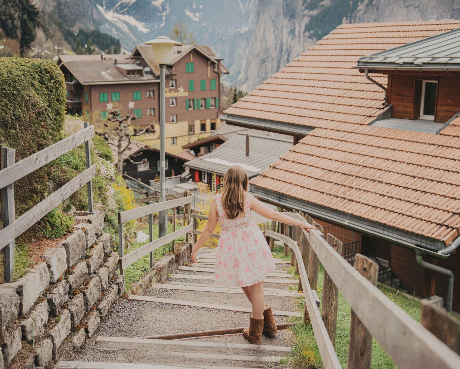 Girl in floral dress walking up stone stairs in Swiss Alpine village with traditional buildings nearby.