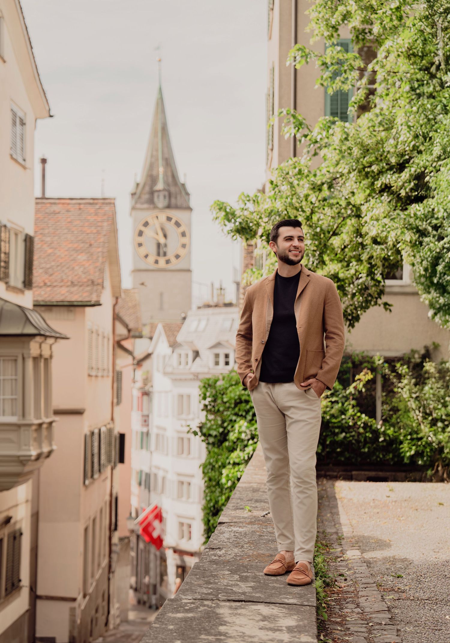 Stylish man in tan blazer and black sweater poses confidently on wall in charming Zurich old town street.