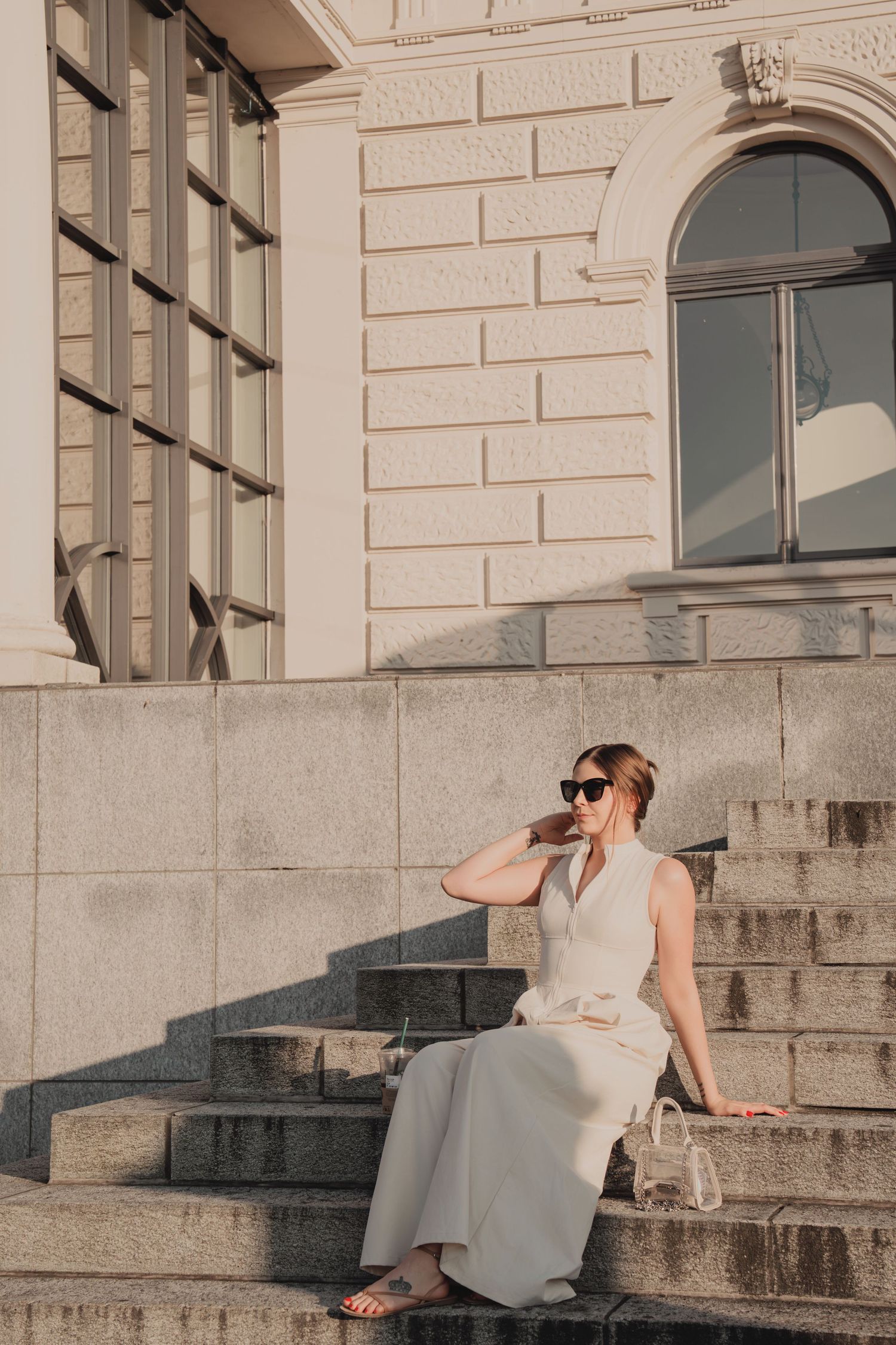 Woman in white sleeveless dress sitting on stone steps outside a classical European building.