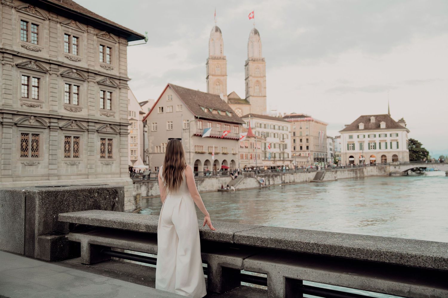 Woman in white dress stands on bridge overlooking Zurich old town with Grossmünster cathedral and Limmat river.