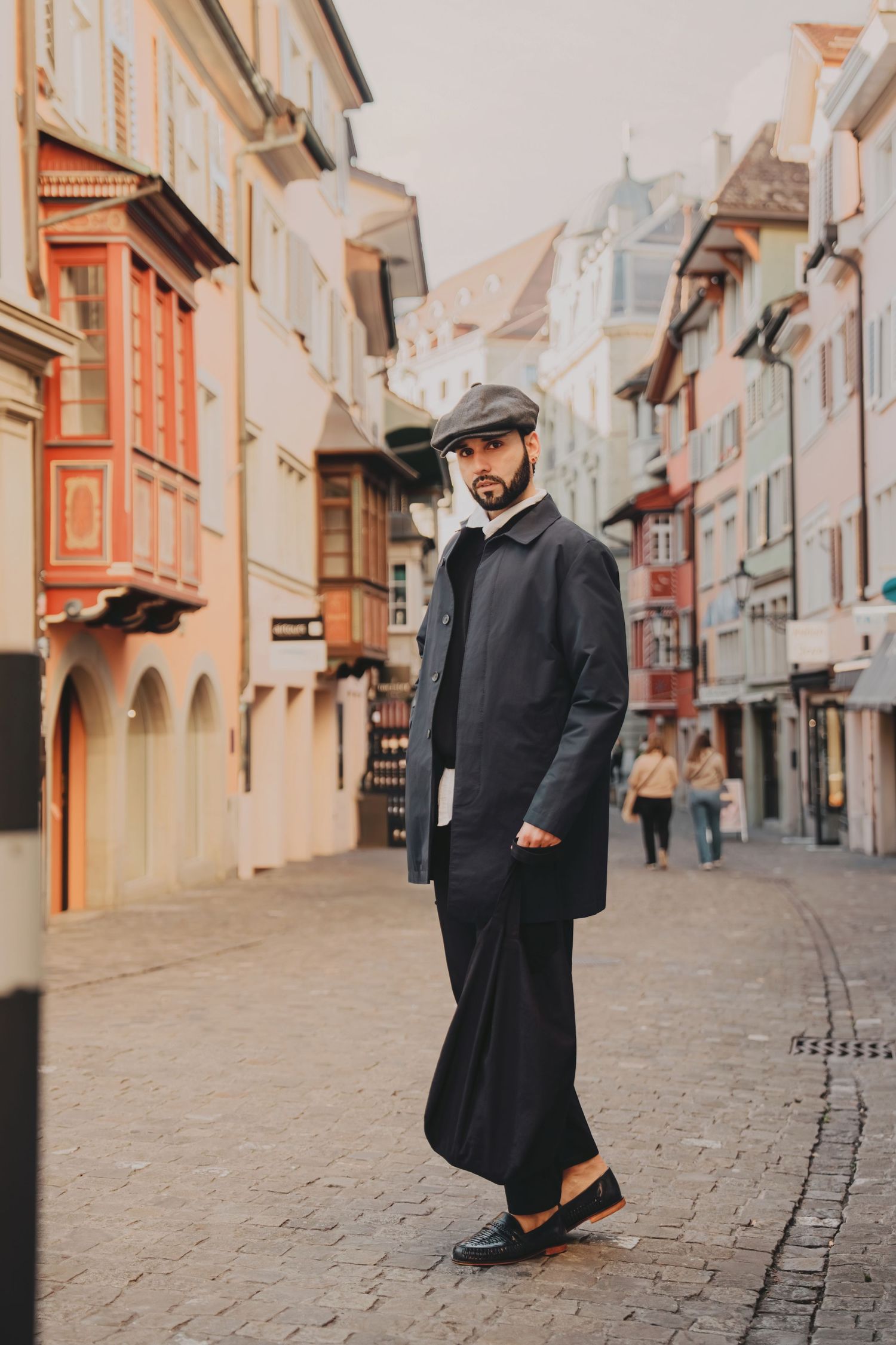 Fashionable bearded man in dark coat and flat cap standing on charming cobblestone European old town street.