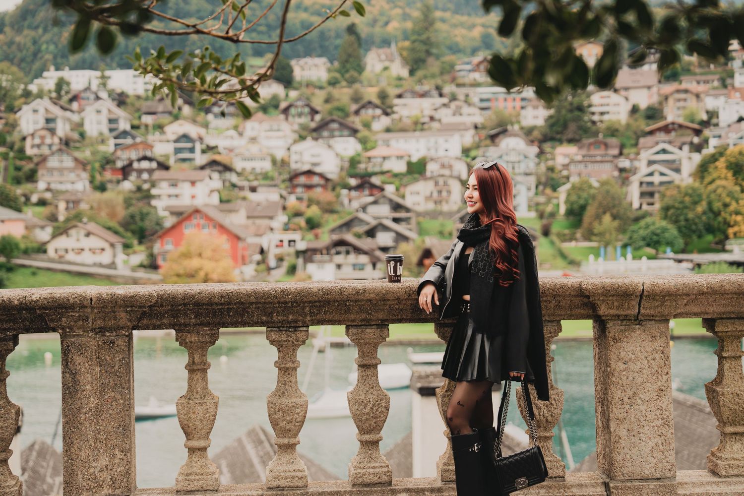 Stylish woman in black cape and leather skirt leans on stone balustrade overlooking Swiss lakeside village.