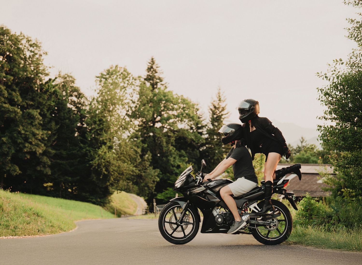 Couple riding together on a black sport motorcycle along a tree-lined road in warm daylight.