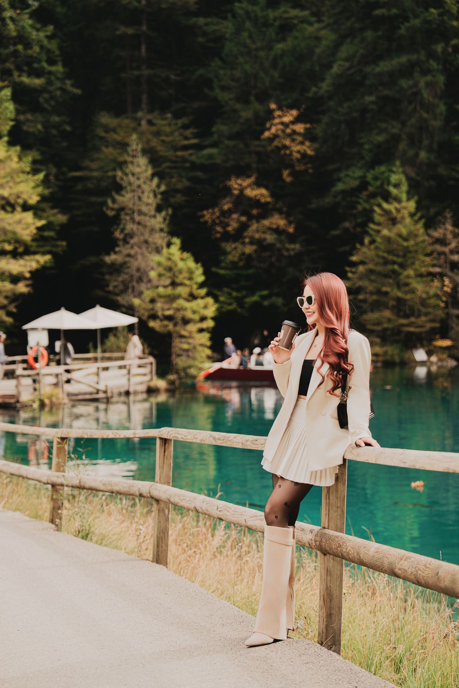 Red-haired woman in white outfit holding coffee cup by turquoise alpine lake with wooden fence.