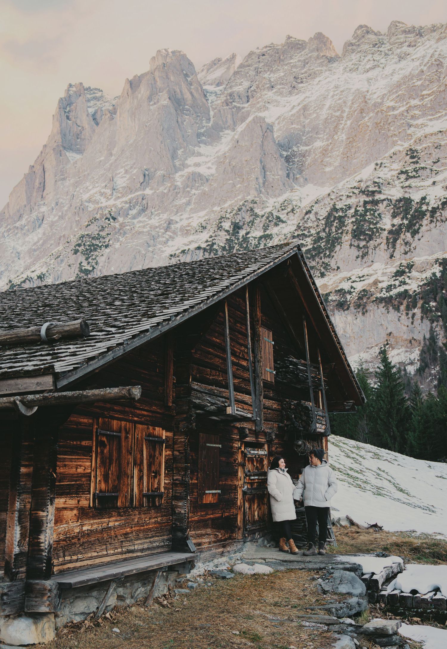 Couple standing together outside rustic wooden alpine cabin with snow-covered rocky mountains behind.