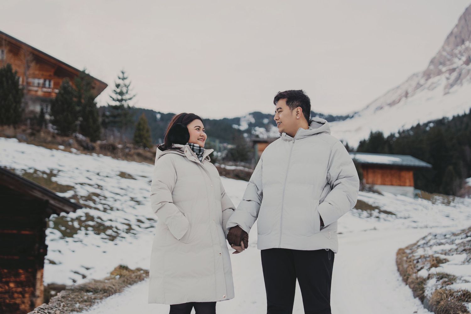 Happy couple holding hands and smiling at each other on snowy path between alpine chalets.
