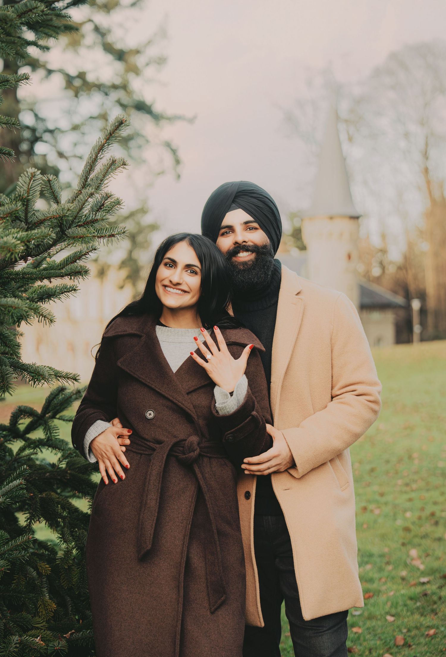 Smiling engaged couple posing near evergreen tree showing off ring at a historic estate.
