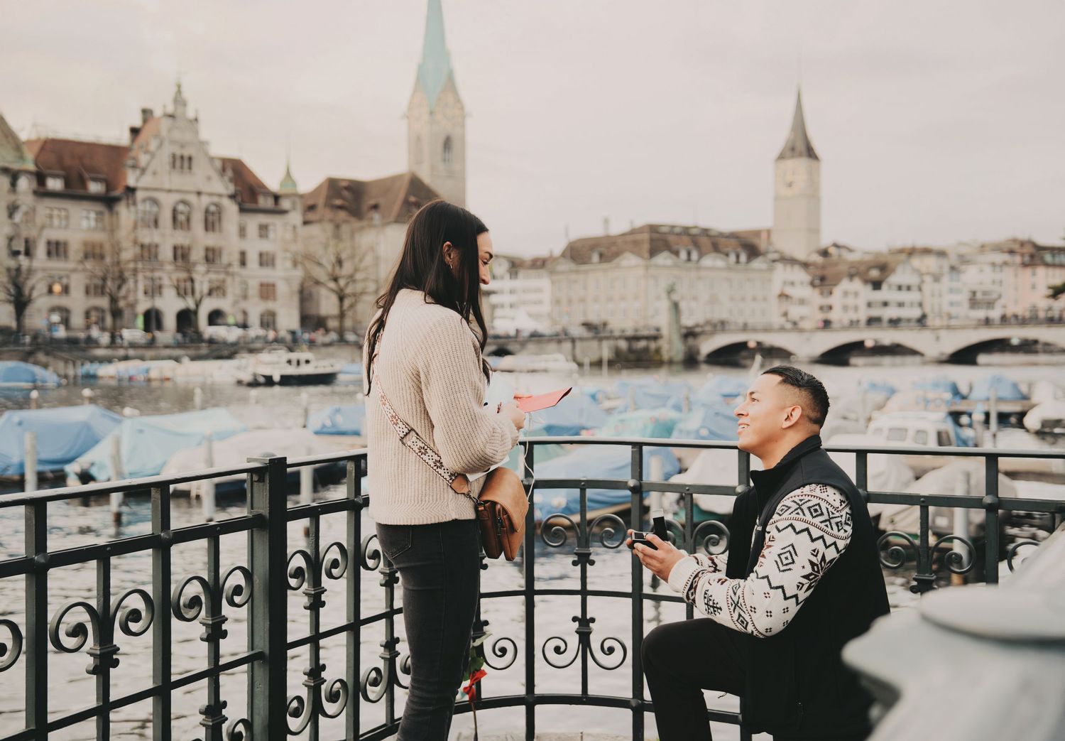 Man proposing on one knee to woman on a bridge overlooking Zurich river and historic skyline.