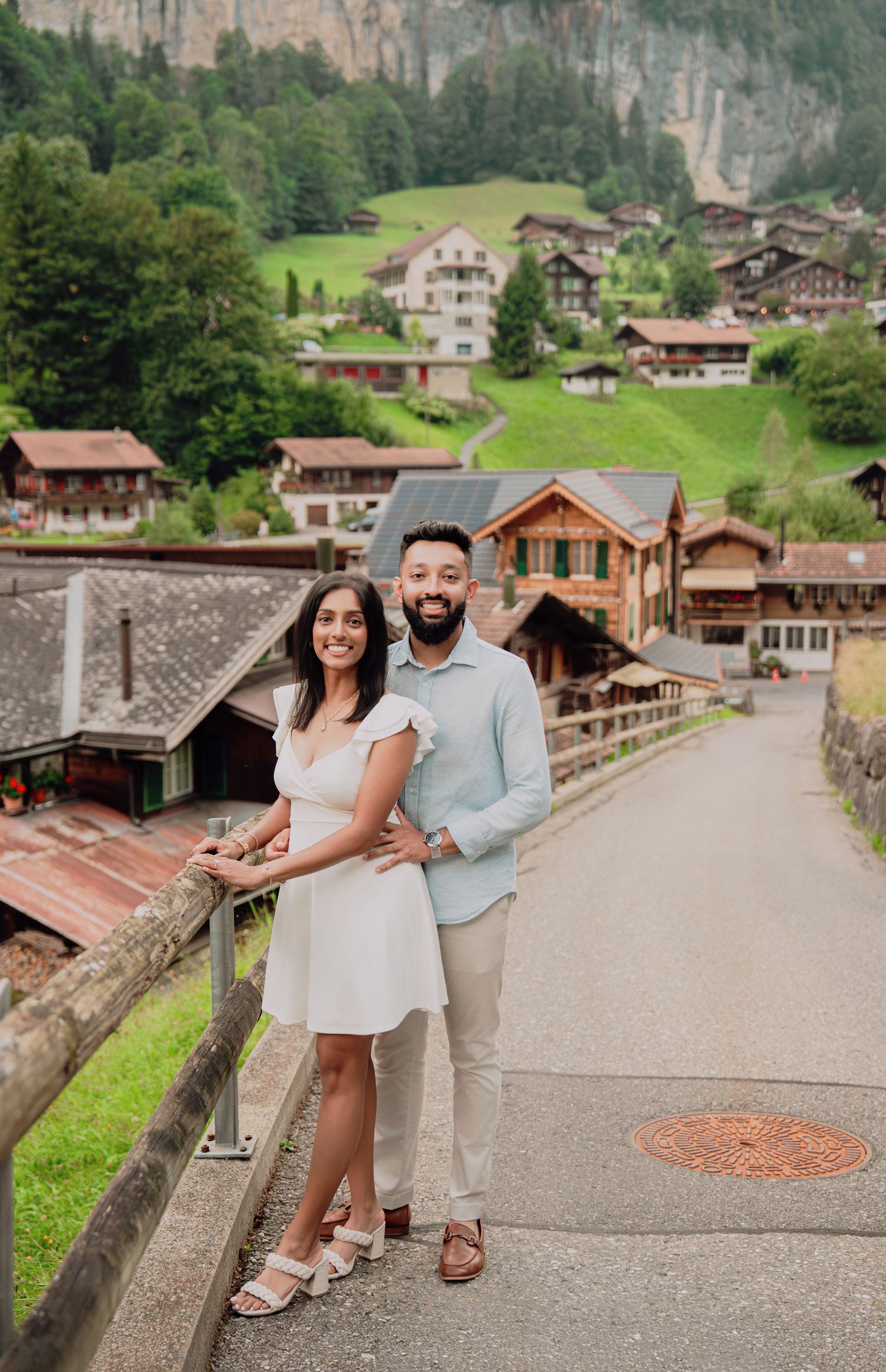 Smiling couple posing on a bridge overlooking a scenic Swiss Alpine village with lush green hillsides.