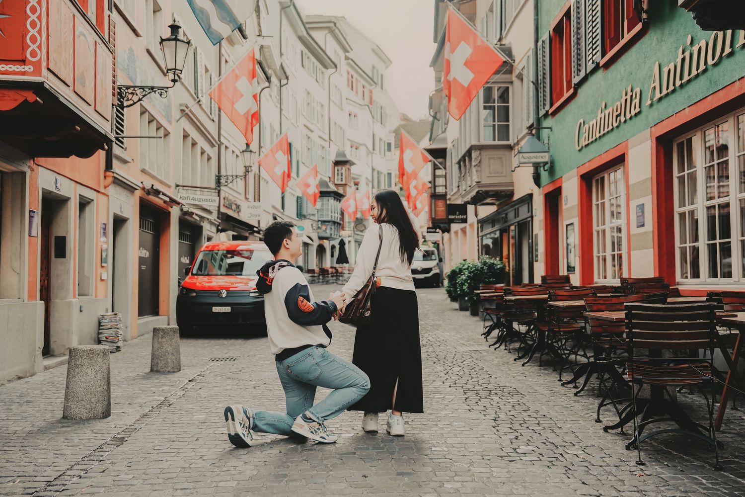 Man proposing on one knee to woman on a cobblestone street in Switzerland with Swiss flags lining the buildings.