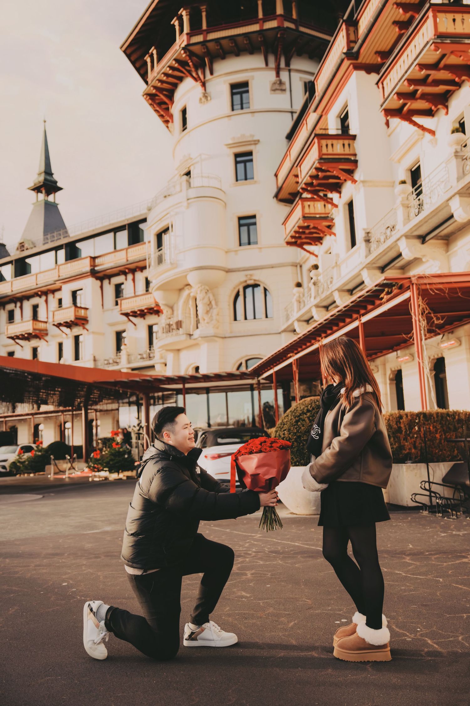 Man on one knee proposing with red roses to his girlfriend outside a grand European luxury hotel.