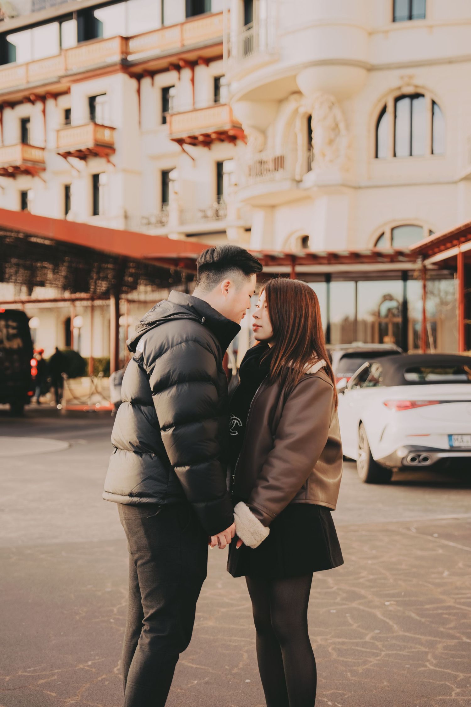 Young Asian couple standing close together romantically on a European city street with buildings behind.
